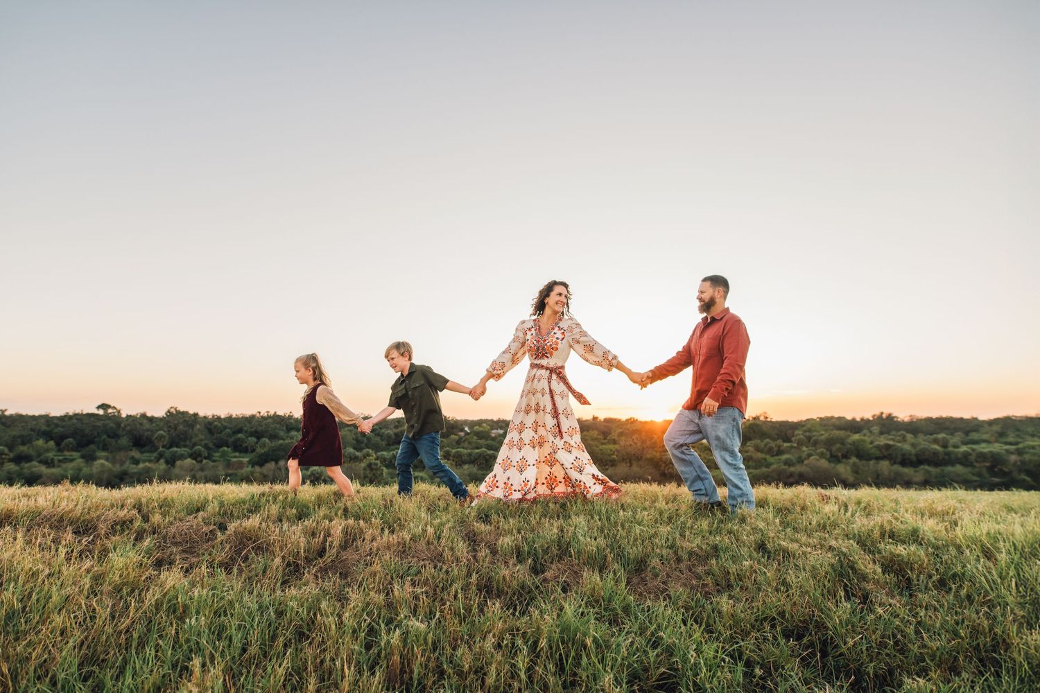 A joyful family holds hands and walks together through a golden field during a beautiful sunset.