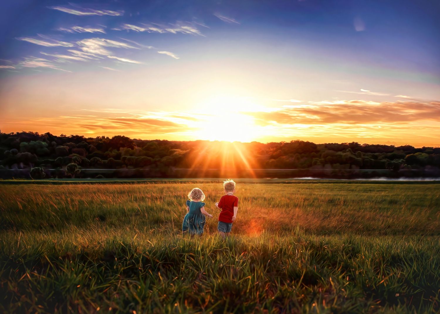 Two children stand in a golden field watching a vibrant sunset over trees.