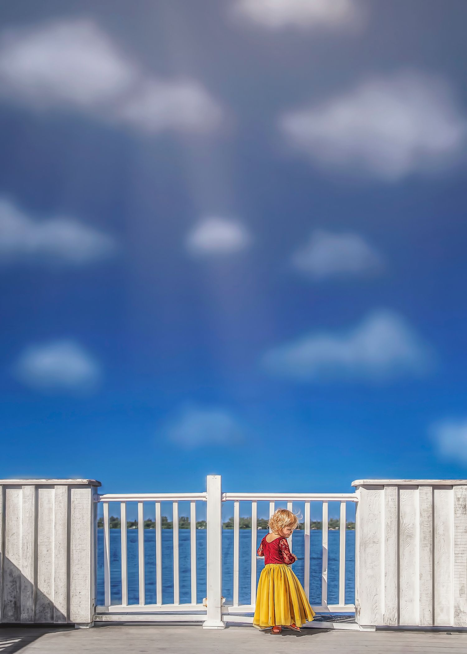 Young girl in red and yellow dress standing on a white railing bridge under blue sky.