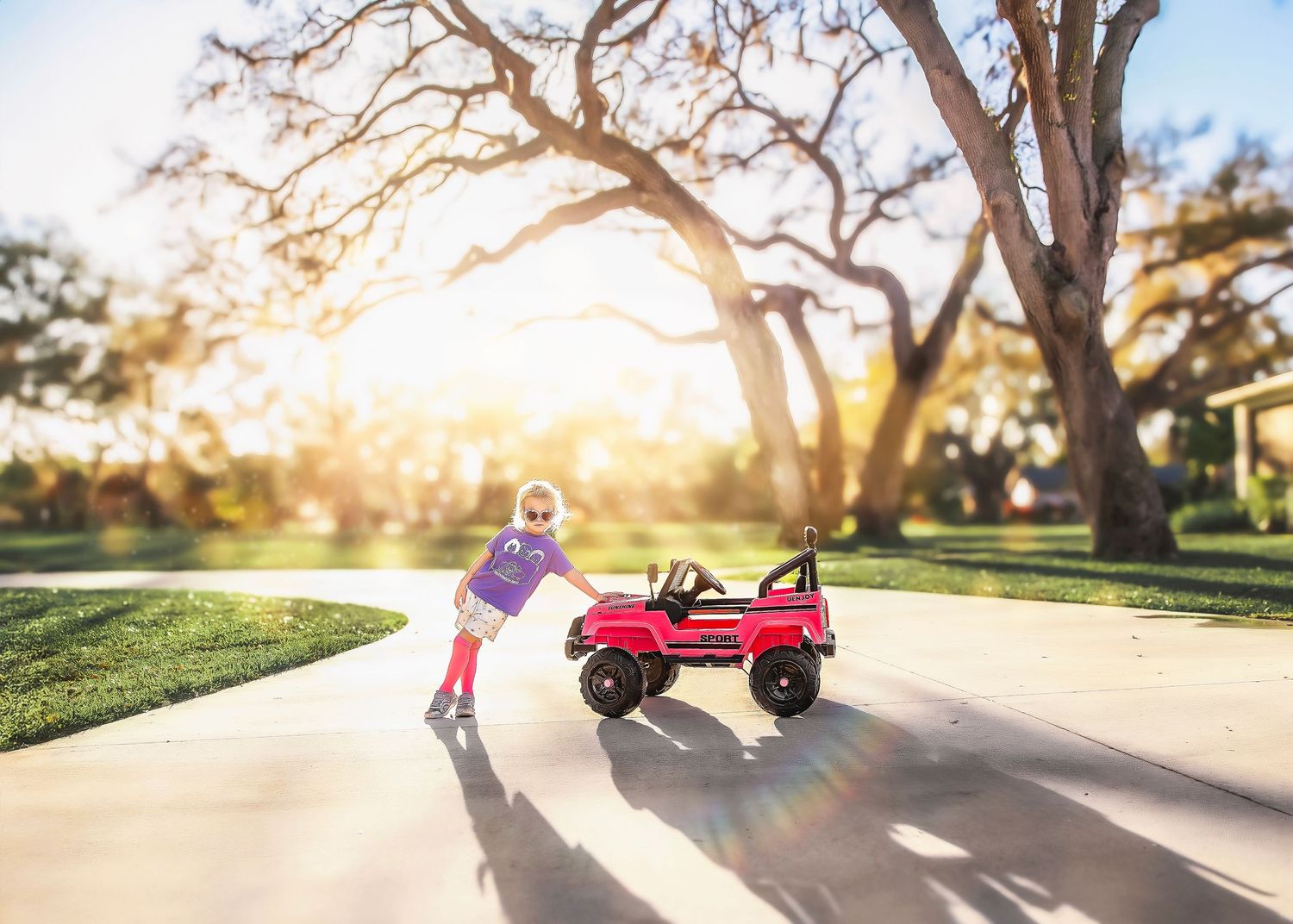 Young child in superhero costume pushing a red toy jeep on a sunny driveway.