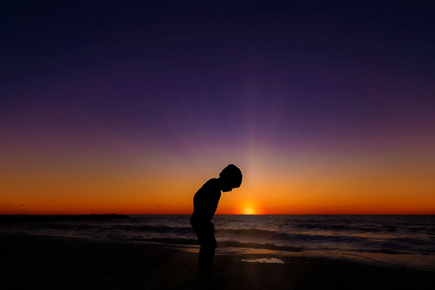 Silhouette of a child standing on a beach during a vibrant orange and purple sunset.