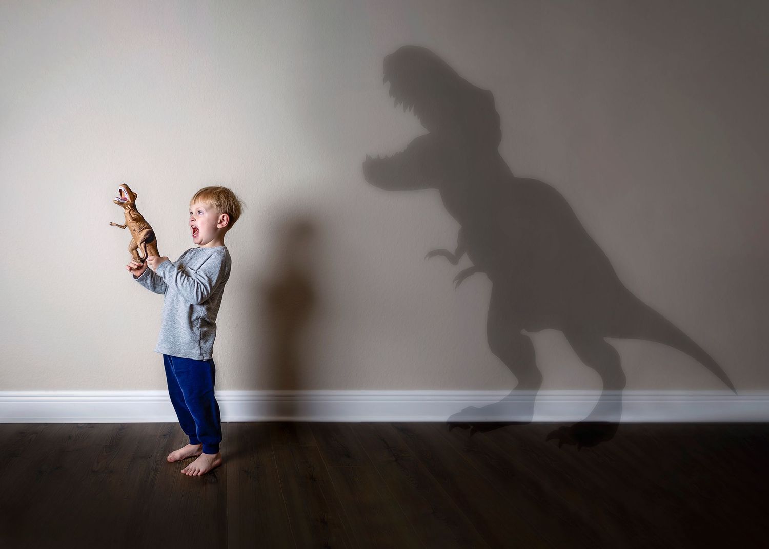 Boy holding toy dinosaur casting a large T-Rex shadow on a white wall indoors.