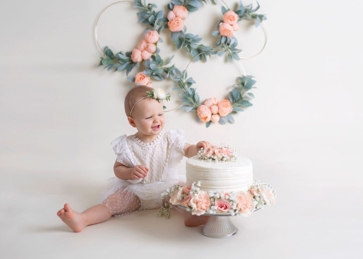 Baby girl in white dress smiling while touching a floral decorated cake at first birthday.