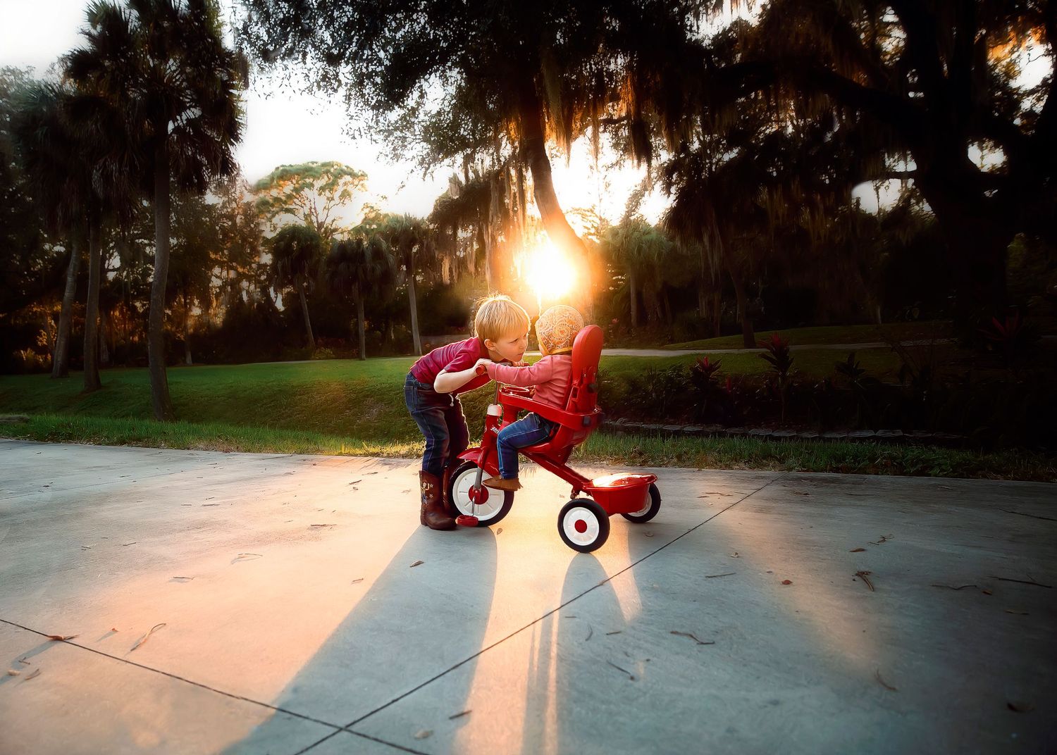Two young children sharing a moment on a red tricycle at sunset with golden sunlight streaming through trees.
