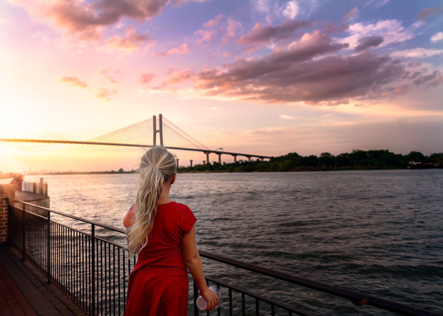 Woman in red dress gazing at a suspension bridge over a river at sunset with colorful pink and purple skies.