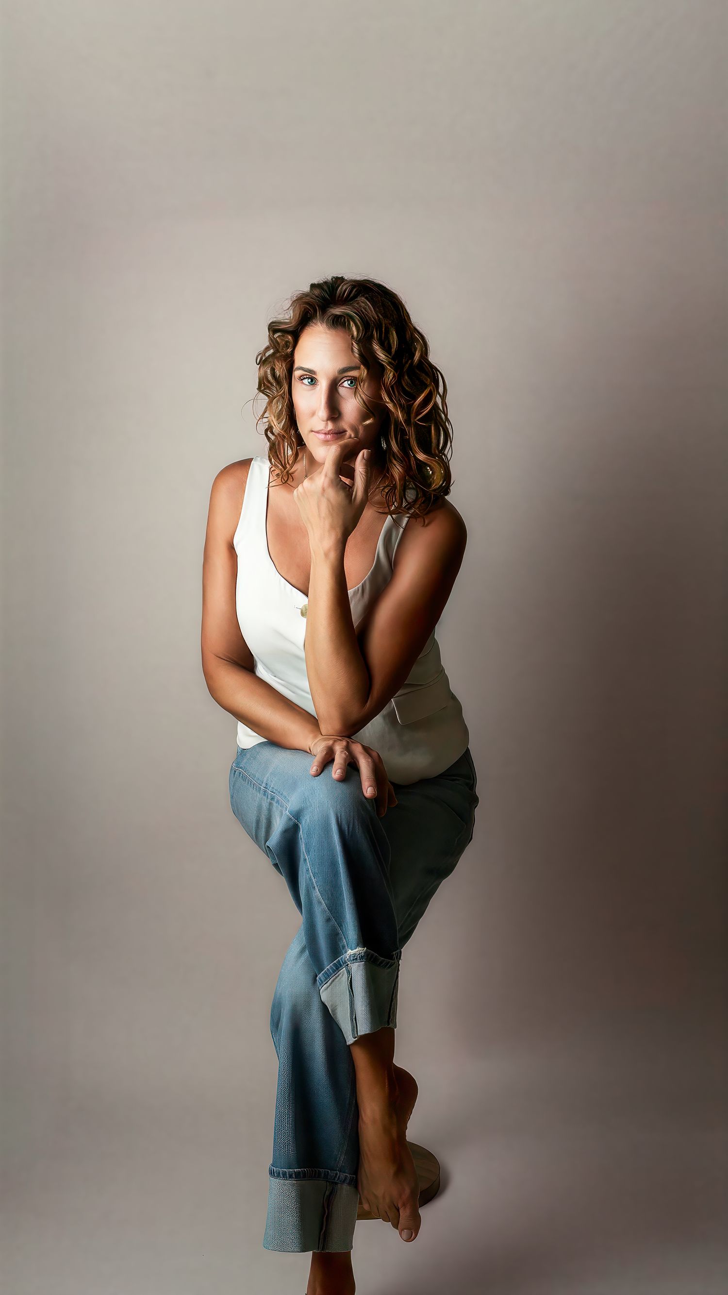 Woman with curly hair in white tank top and jeans sits barefoot against neutral gray background.