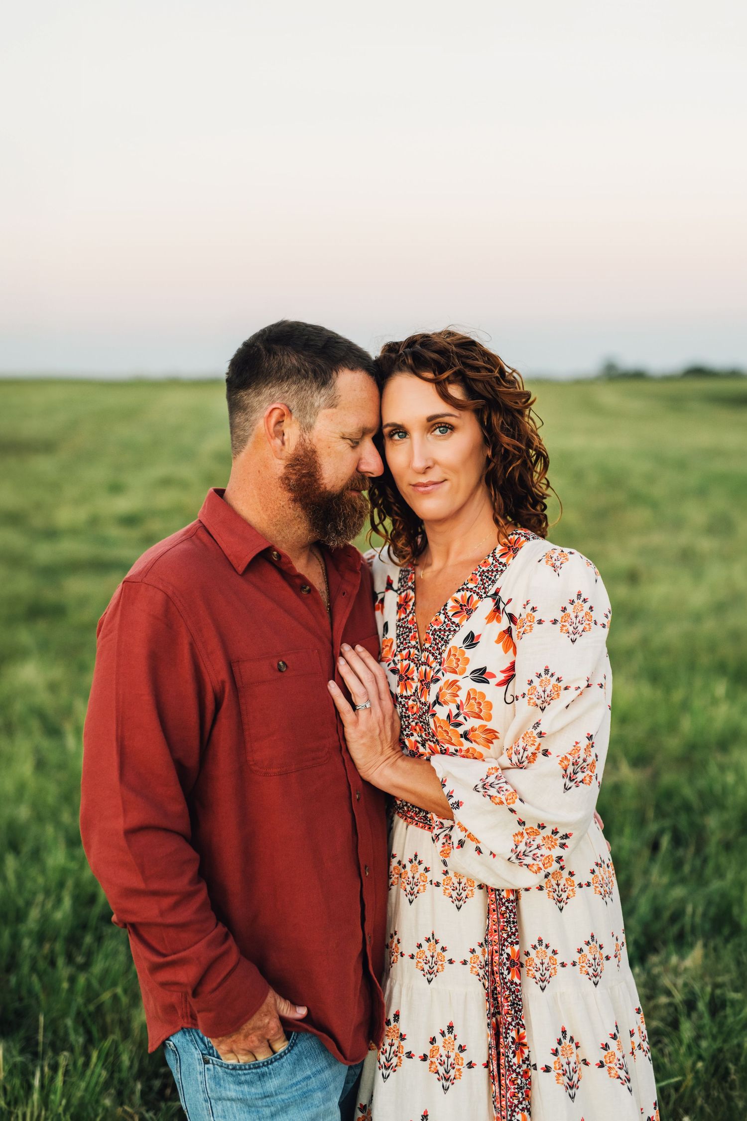 A couple embraces tenderly in a green field at sunset, the man in red and woman in a floral dress.