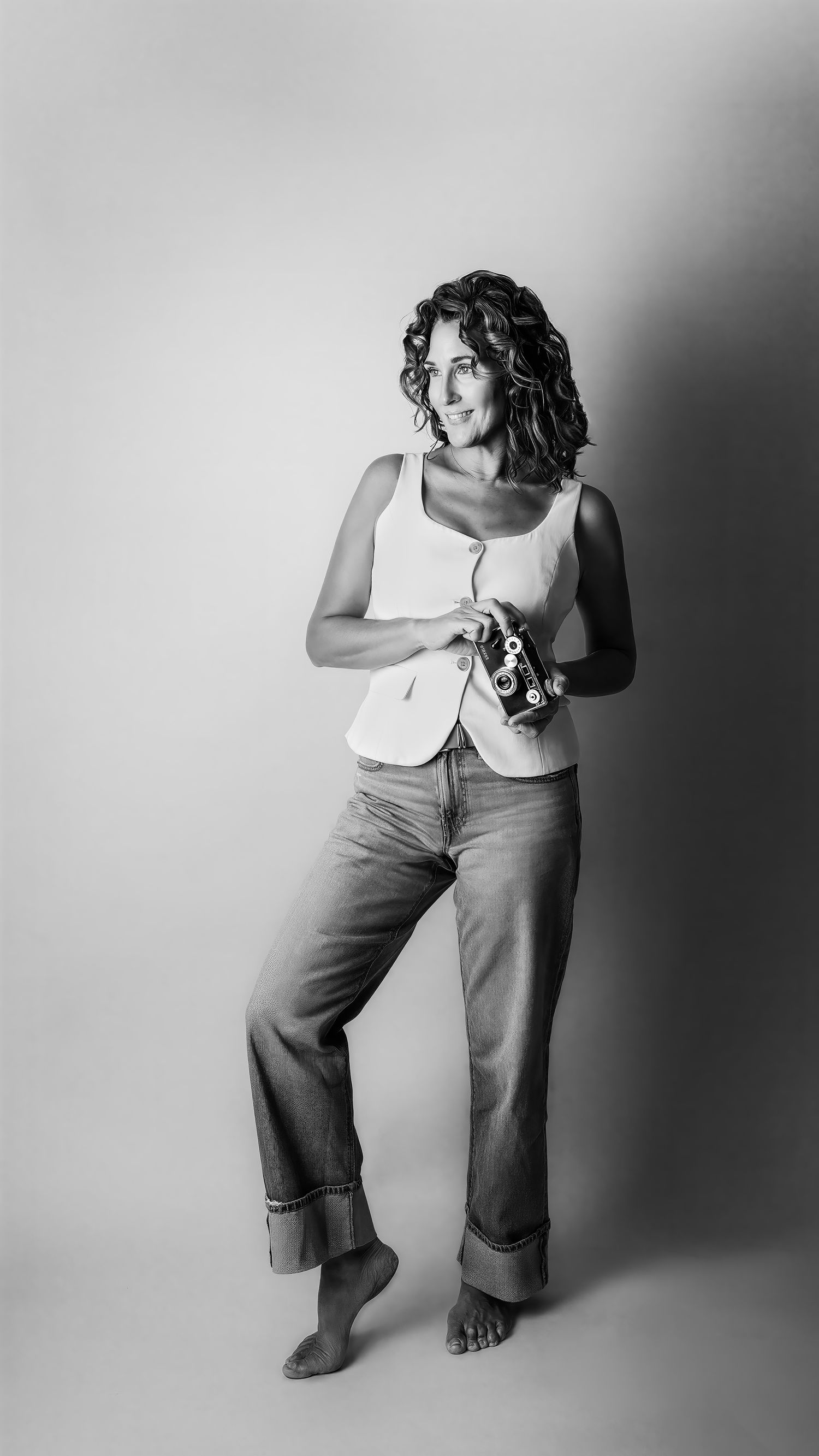 Black and white portrait of a curly-haired woman in white top and wide-leg jeans holding a vintage camera.