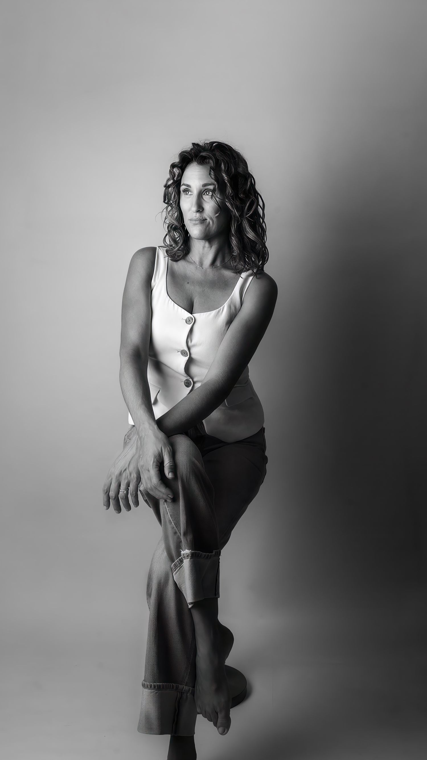 Black and white studio portrait of a woman with curly hair in a white tank top and jeans, posing confidently.