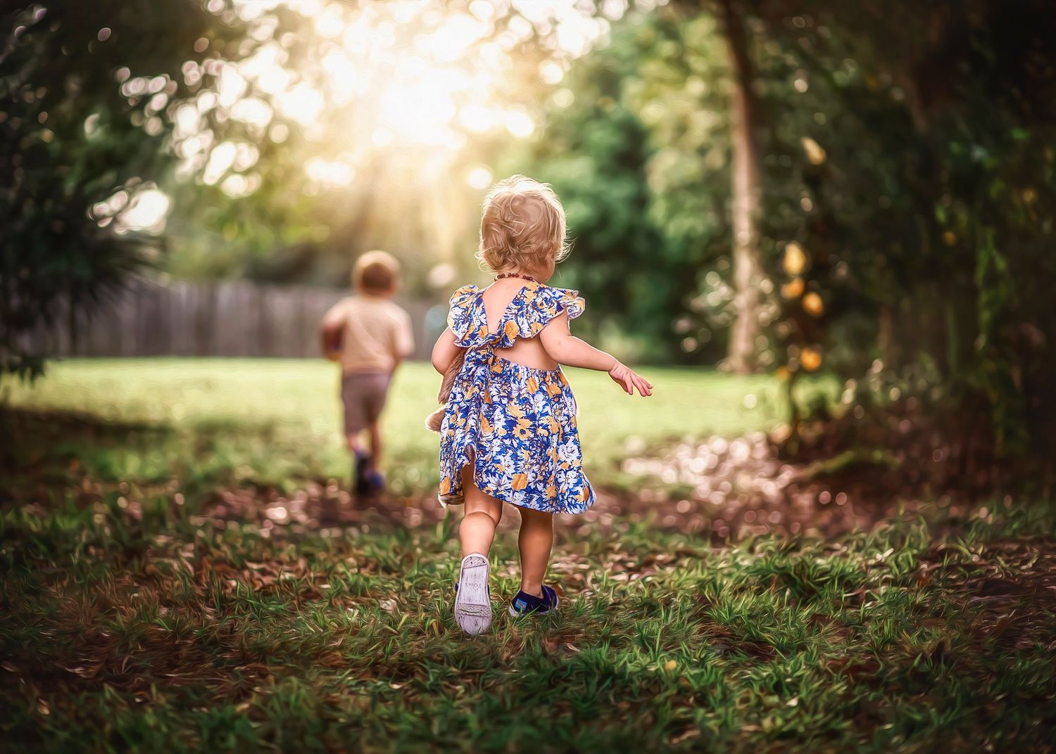 Two young children running through a sunlit garden path surrounded by lush green trees.