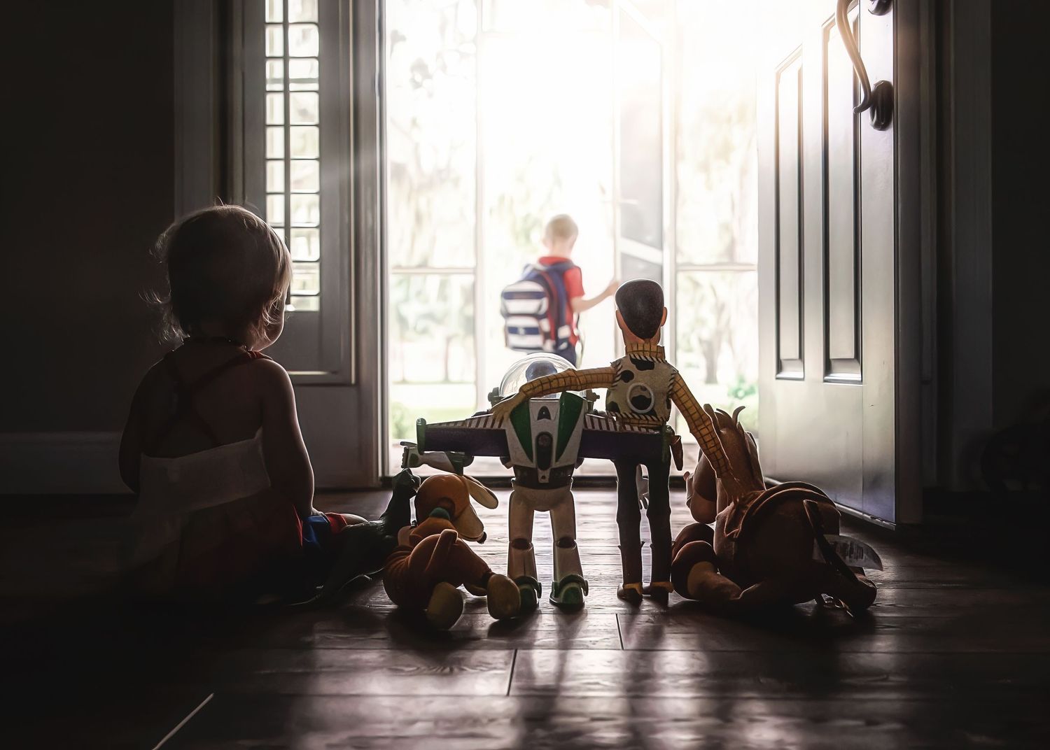 Children playing with toys on a dark hallway floor, silhouetted against bright light from an open front door.