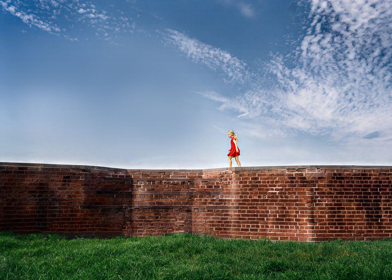 Young girl in a red dress standing atop a curved historic brick wall under a blue cloudy sky.