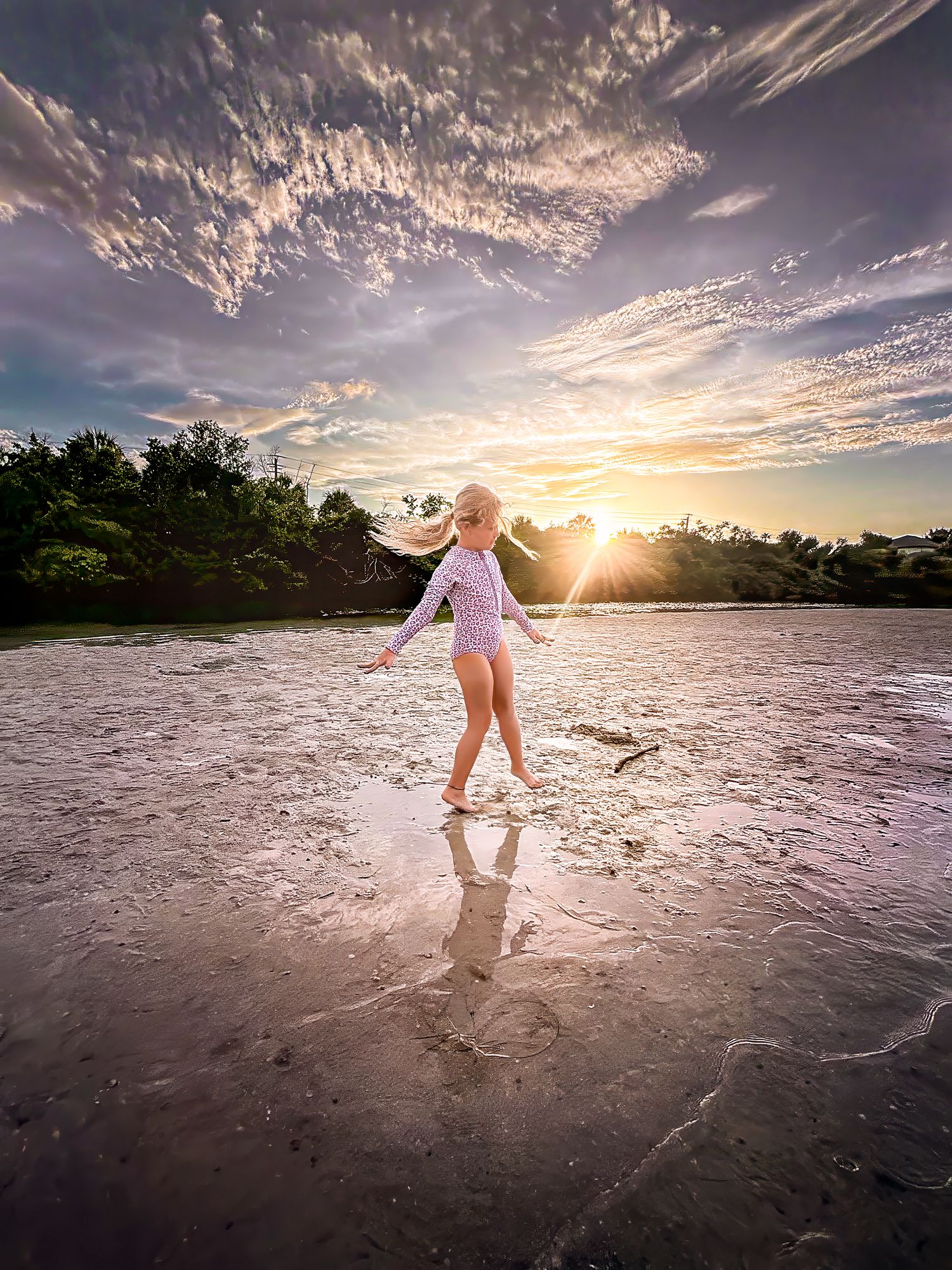 A blonde girl in a swimsuit dances on a wet beach with a dramatic golden sunset and cloudy sky.