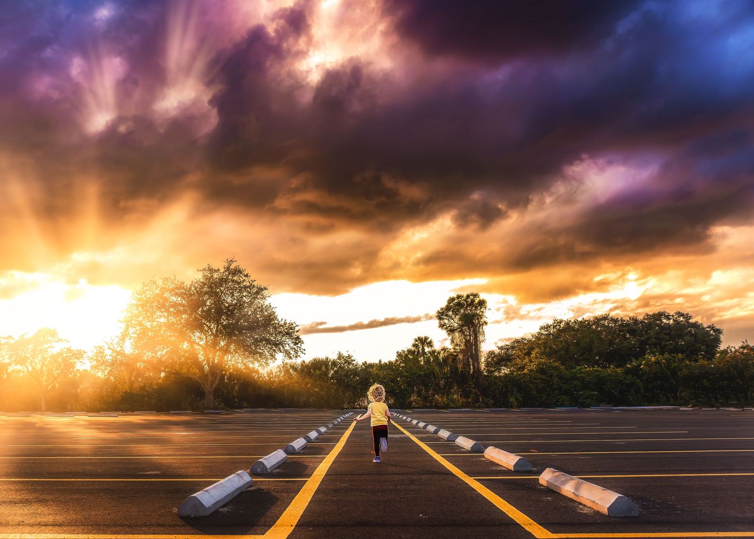 Child running toward a dramatic sunset on an empty road lined with trees.