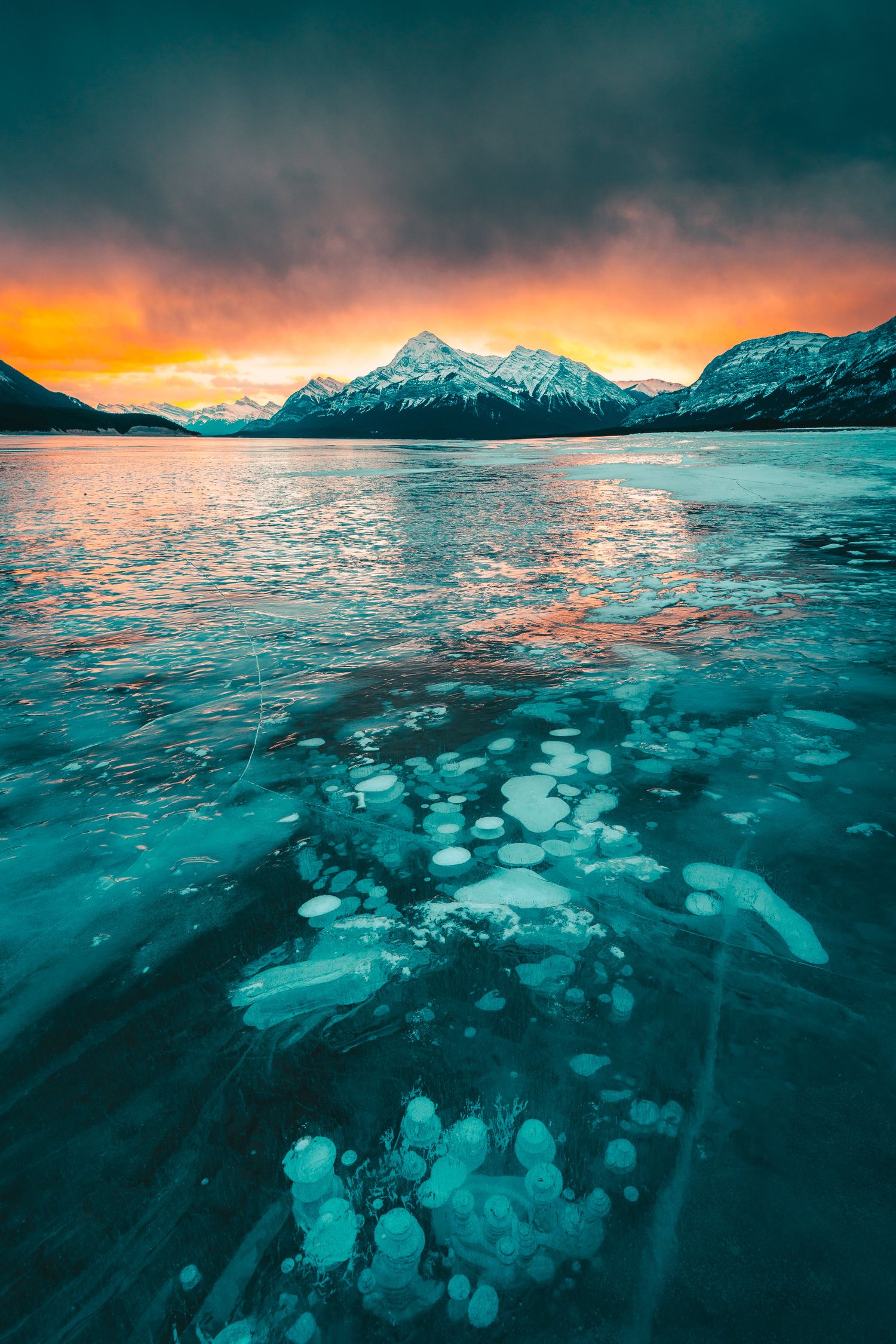 Frozen lake with methane bubbles beneath ice at sunset with snow-capped mountains and dramatic sky.