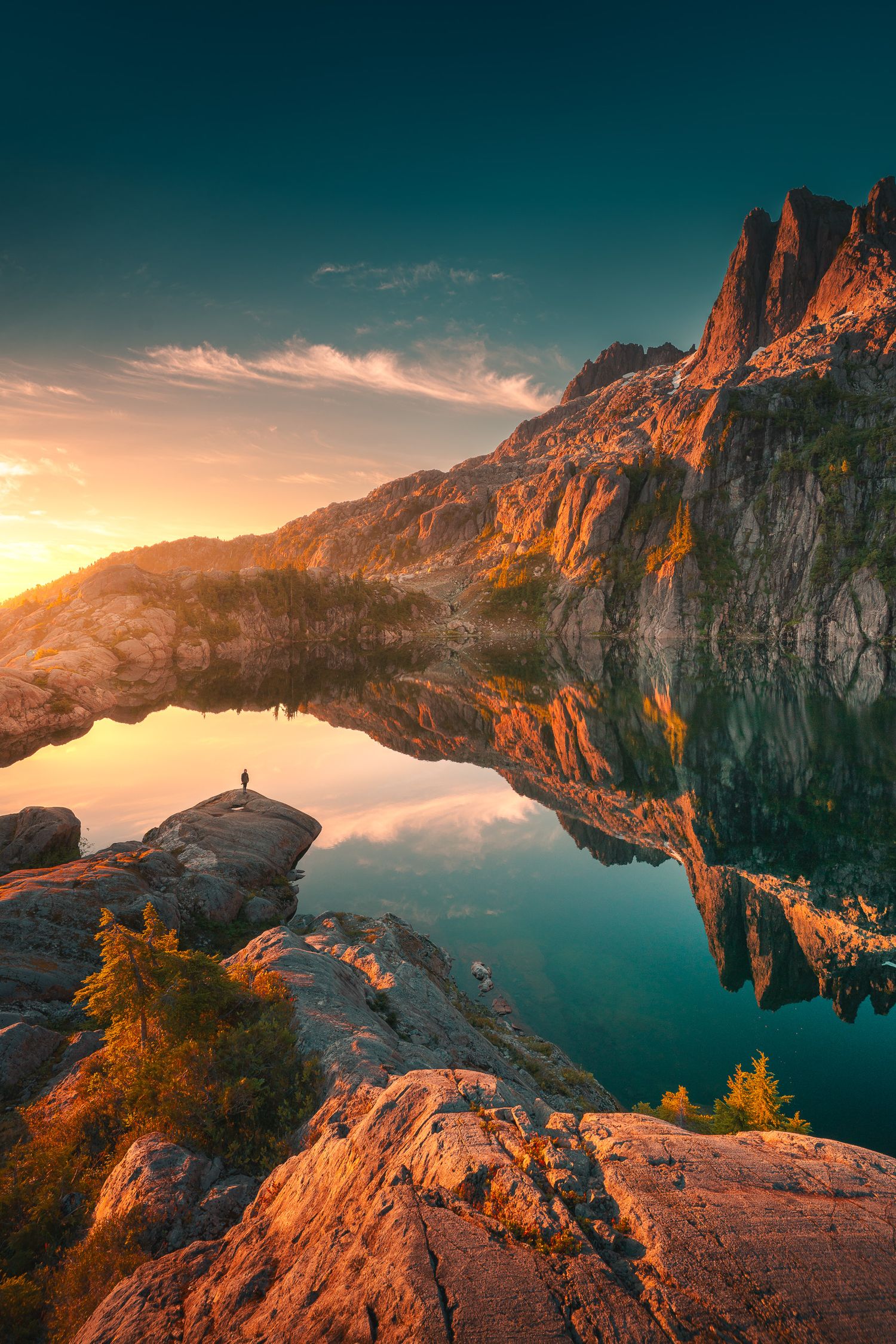 A lone hiker stands on rocky shores of a mirror-like alpine lake reflecting golden sunset and dramatic mountain peaks.