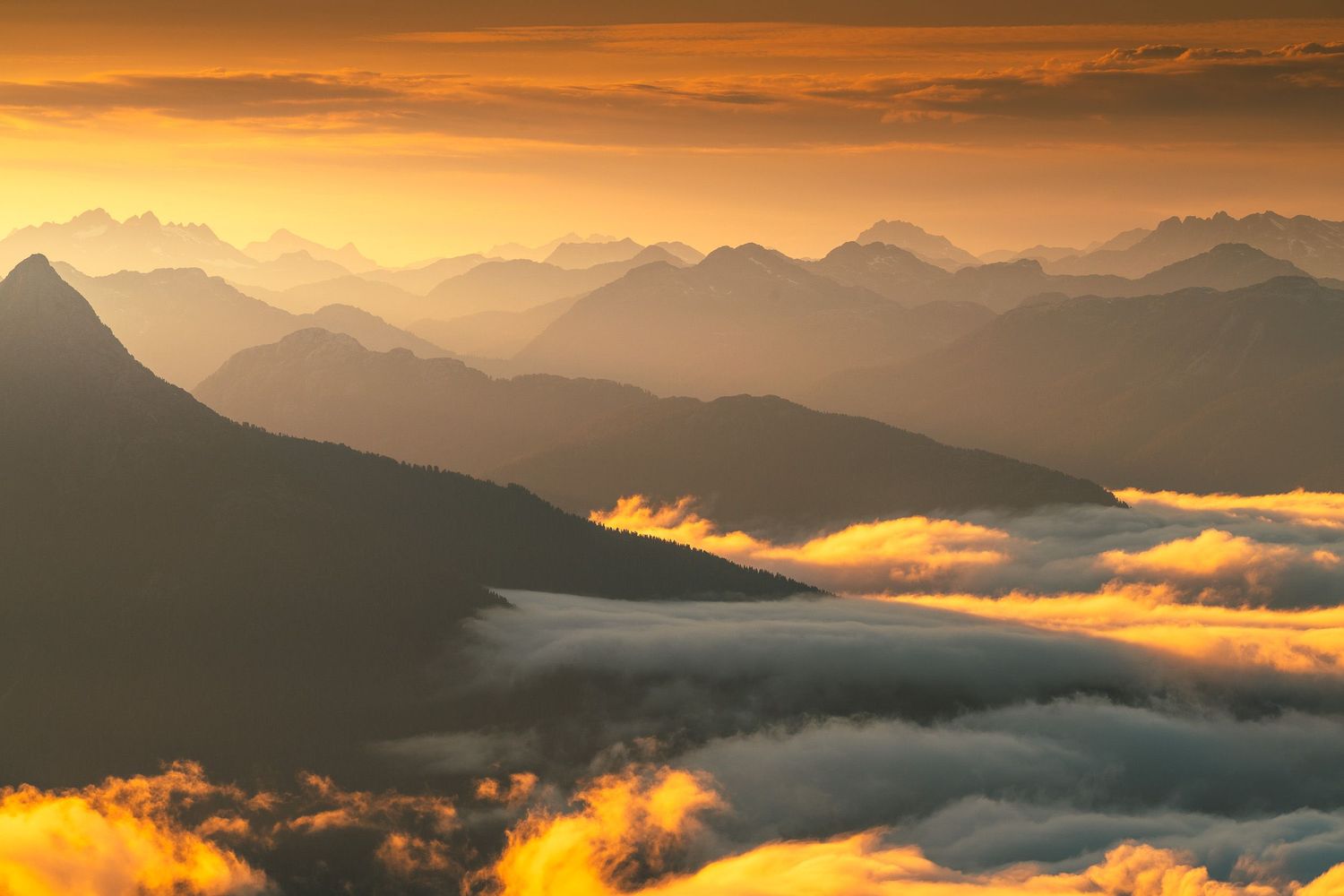 Golden sunset over layered mountain silhouettes with dramatic clouds and mist-filled valleys below.