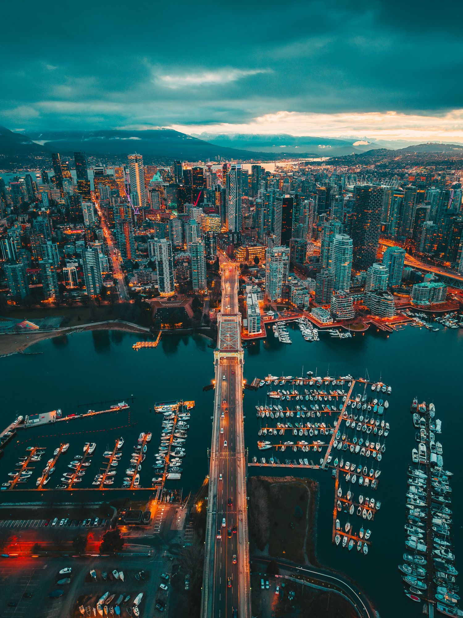 Aerial twilight view of Vancouver city skyline with glowing bridge spanning a marina-lined waterway.