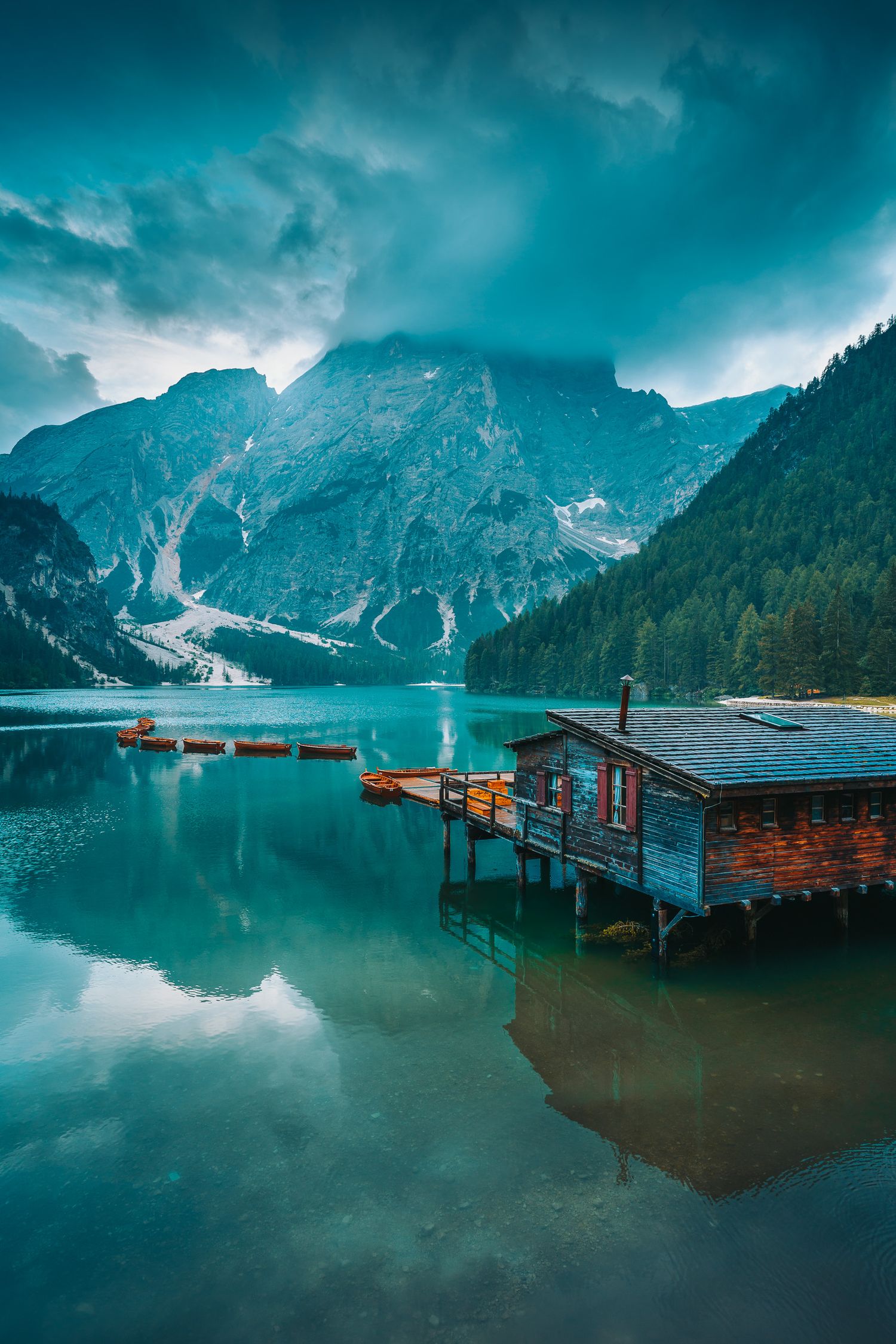 Rustic wooden boathouse on a turquoise alpine lake with dramatic mountain reflections under stormy skies.