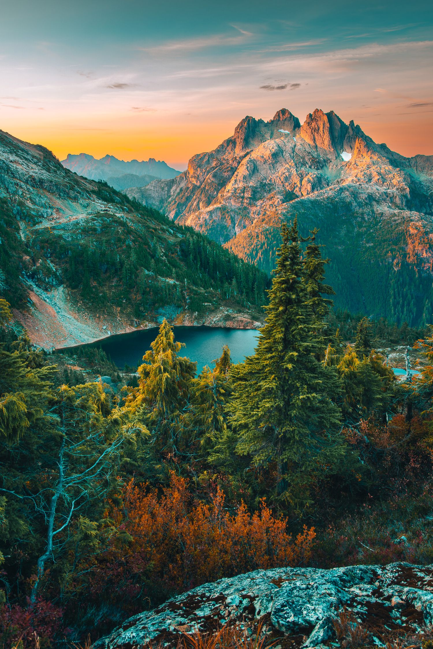 Alpine lake surrounded by autumn forest and rocky peaks glowing at sunset.