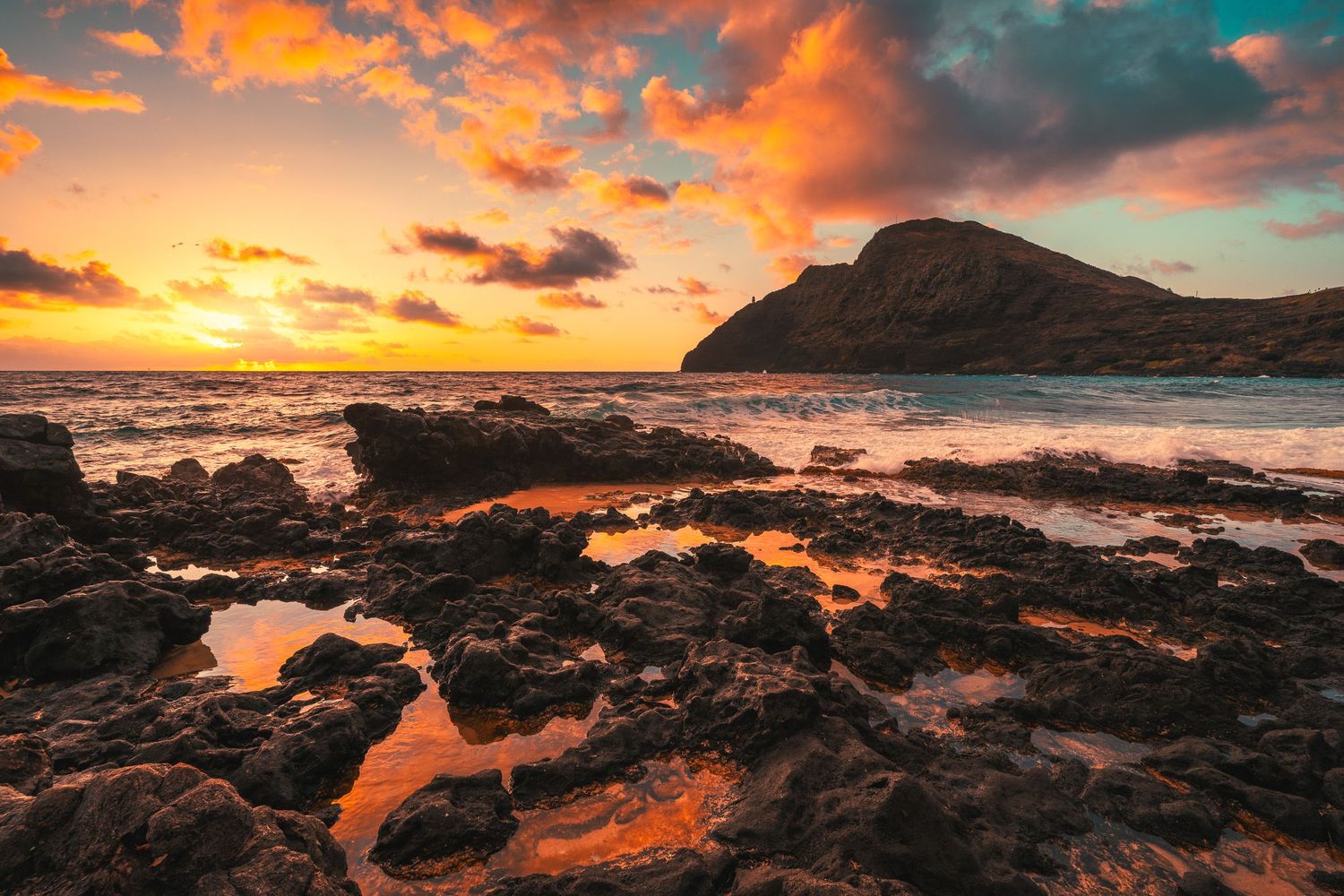 Vibrant sunset over rocky Hawaiian coastline with tide pools reflecting orange and red sky.