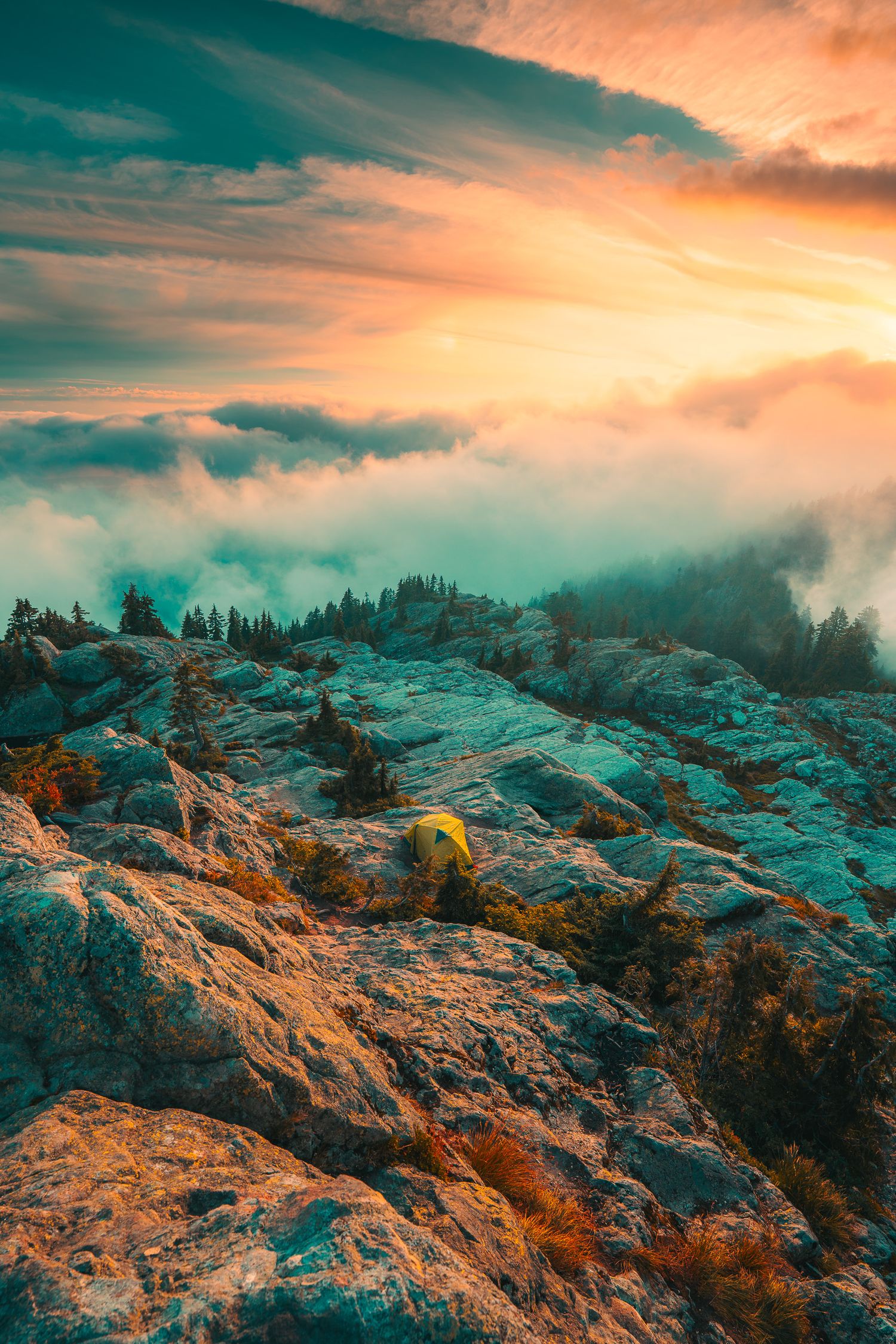 Yellow camping tent on rocky mountain summit above a sea of clouds at a colorful sunset.