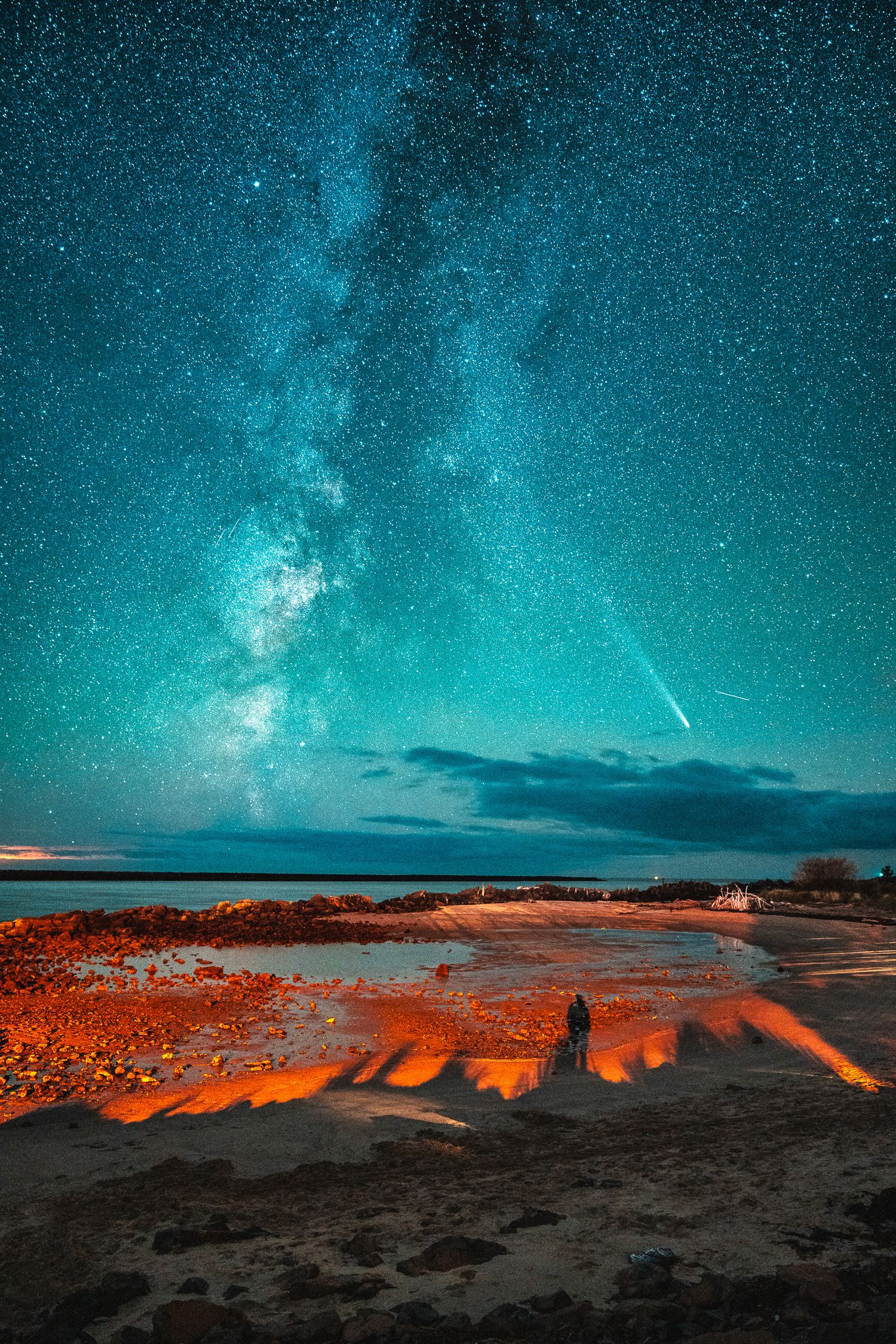 A lone figure stands on a sandy beach under a vibrant Milky Way with a comet streaking across the sky.