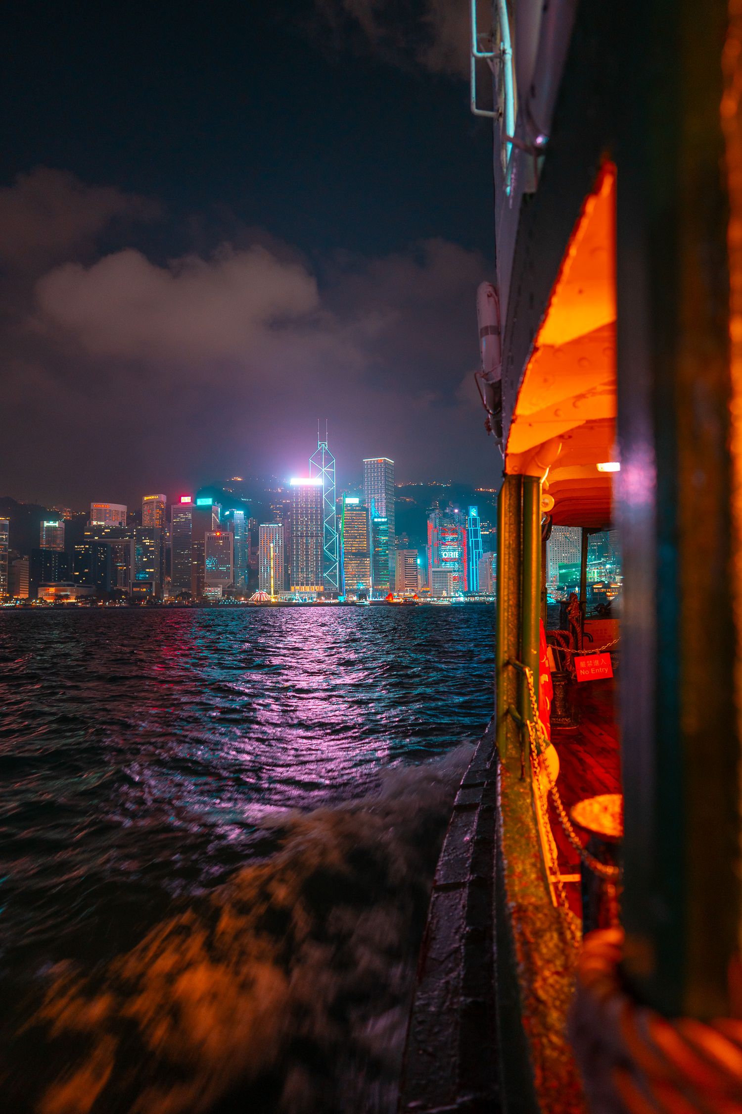 A ferry boat crosses Victoria Harbour at night with Hong Kong's glittering neon skyline reflected on dark waters.