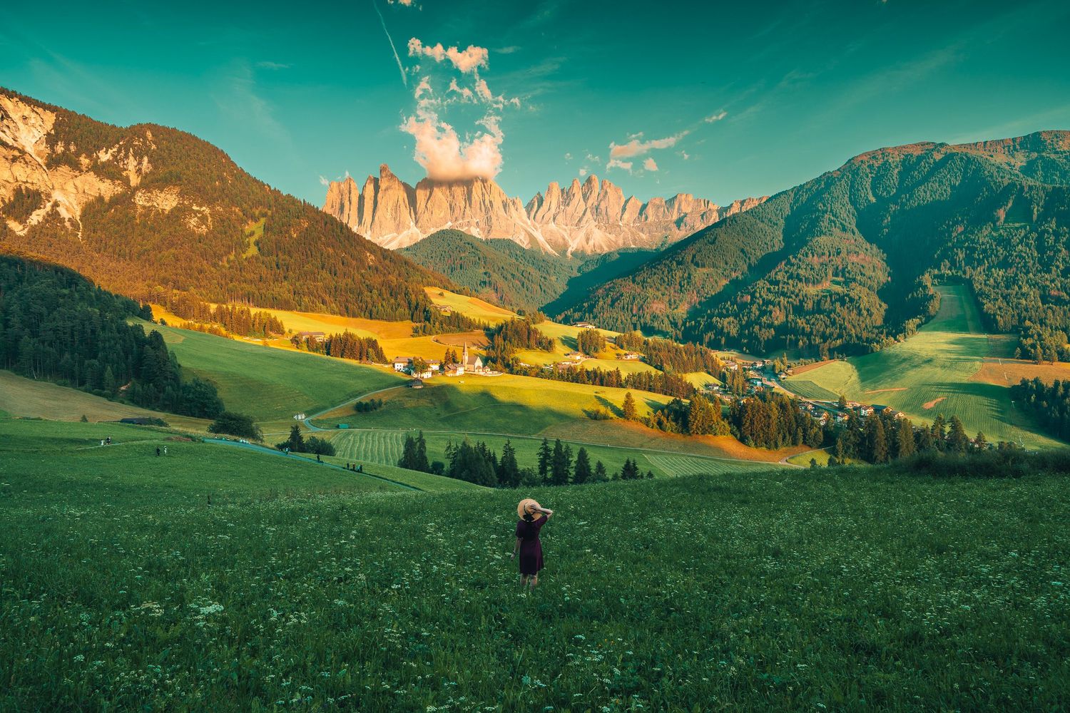 Person standing in lush green valley meadow with Dolomite mountains and village backdrop.