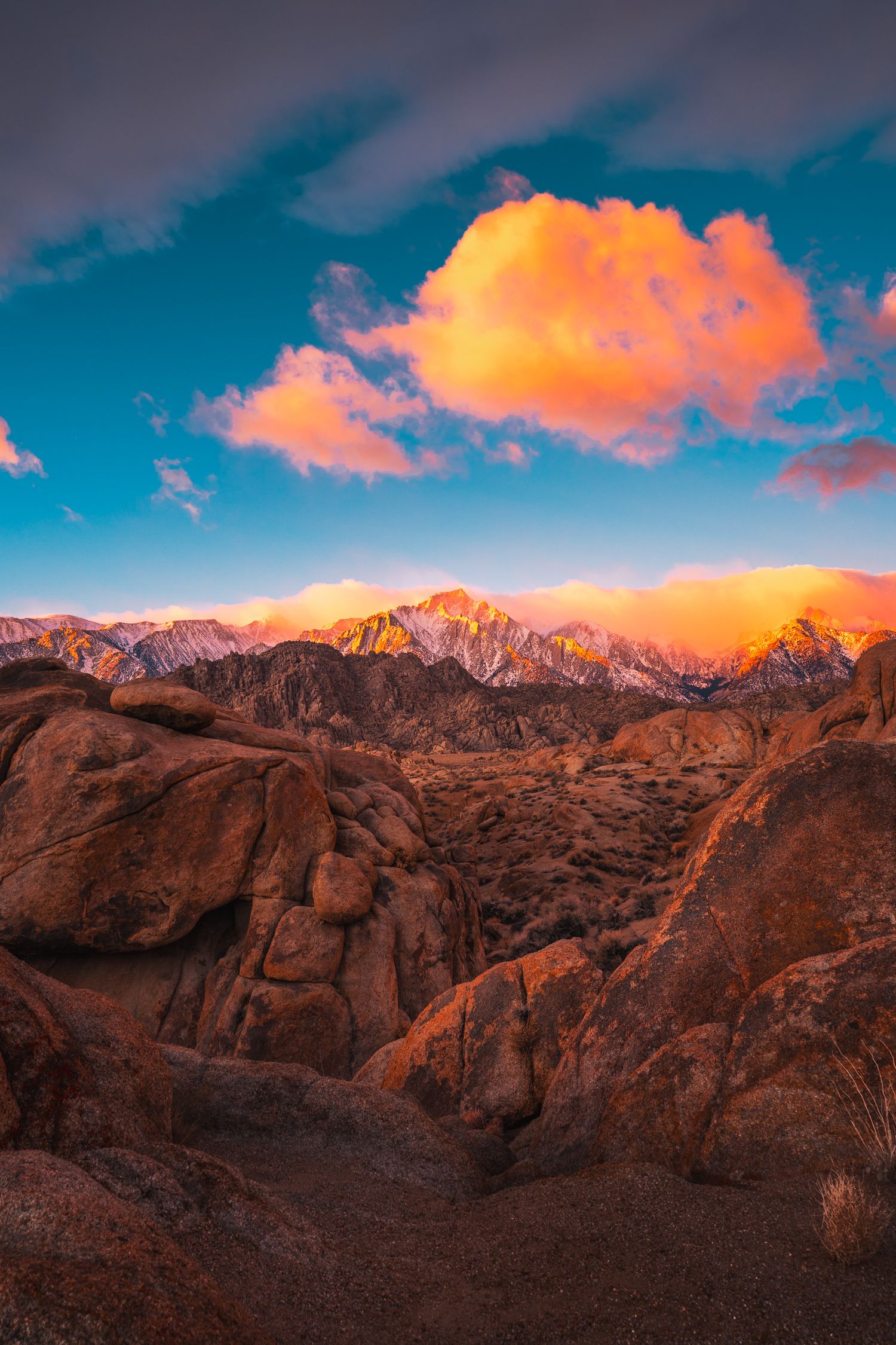 Golden sunset illuminates red rock boulders and mountain peaks under a vivid blue sky with orange clouds.