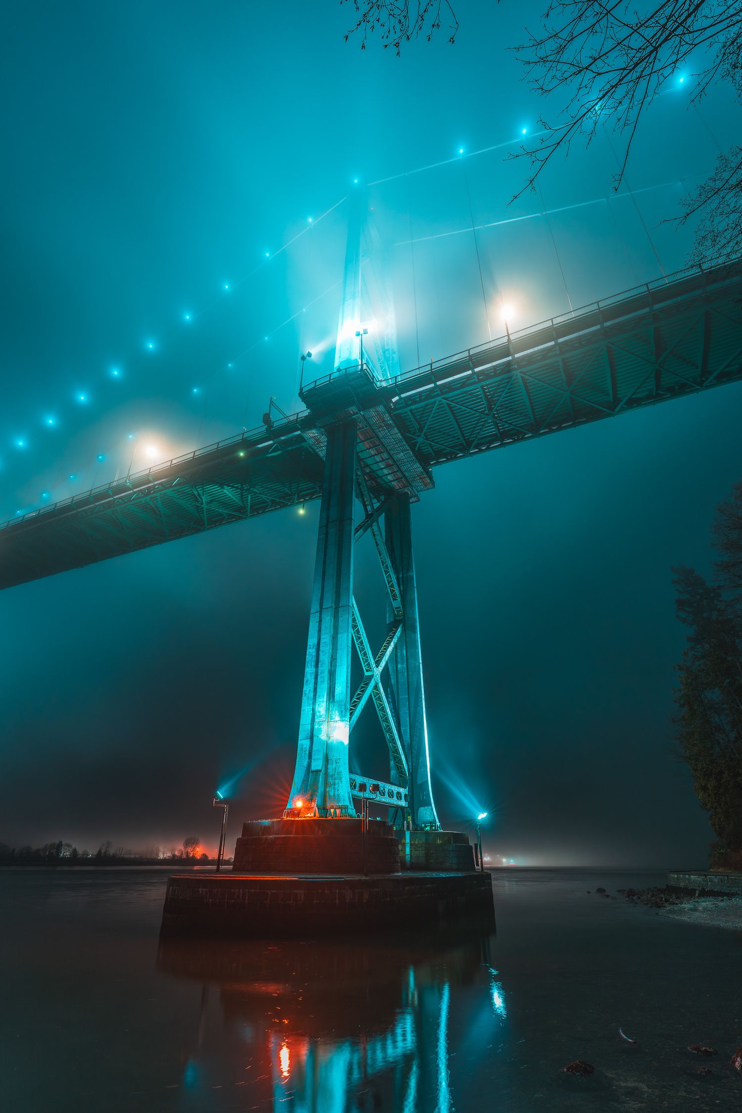 Lions Gate Bridge tower illuminated in teal light emerges dramatically through dense fog over calm dark waters.
