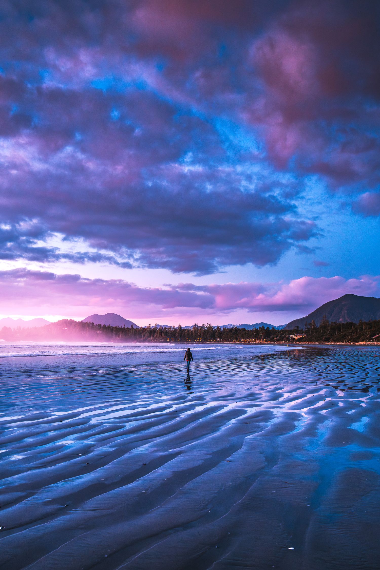 A solitary figure stands on a rippled tidal flat beneath dramatic purple and pink sunset clouds.