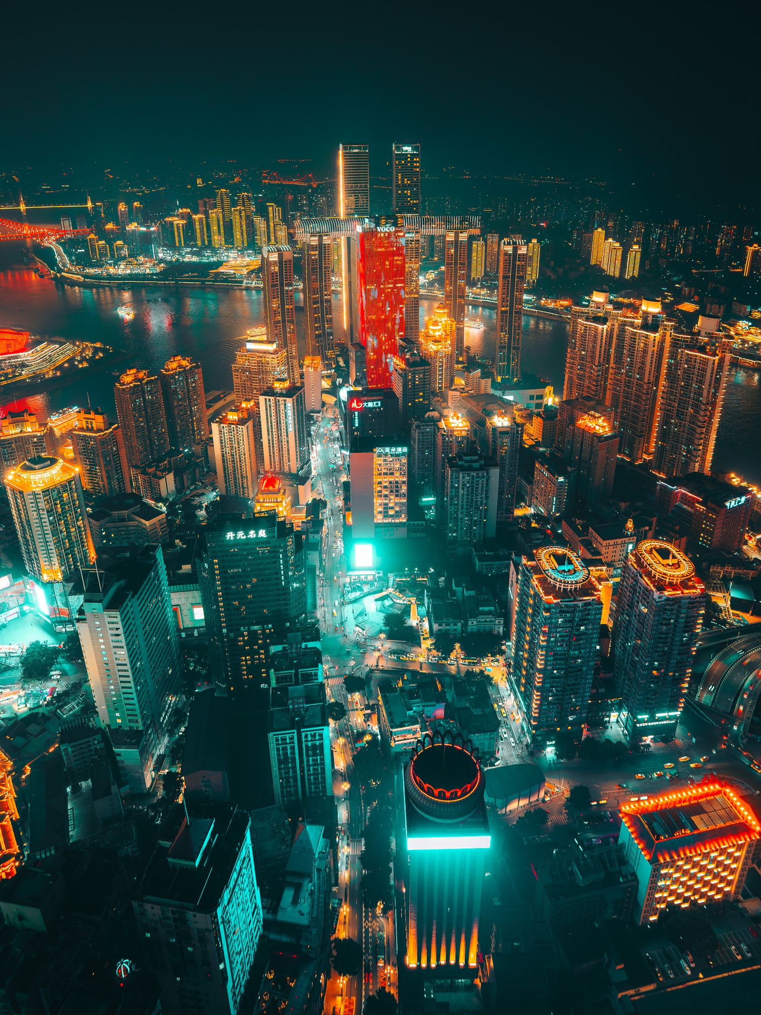 Vibrant aerial night panorama of Chongqing's dense urban skyline with teal and orange neon lights by the river.