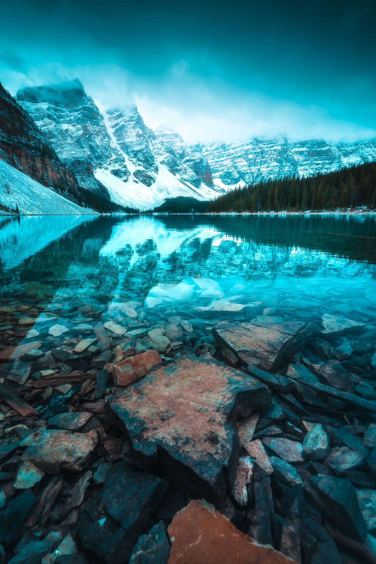 Crystal-clear turquoise glacial lake reflecting snow-capped mountains with rocky shoreline foreground.