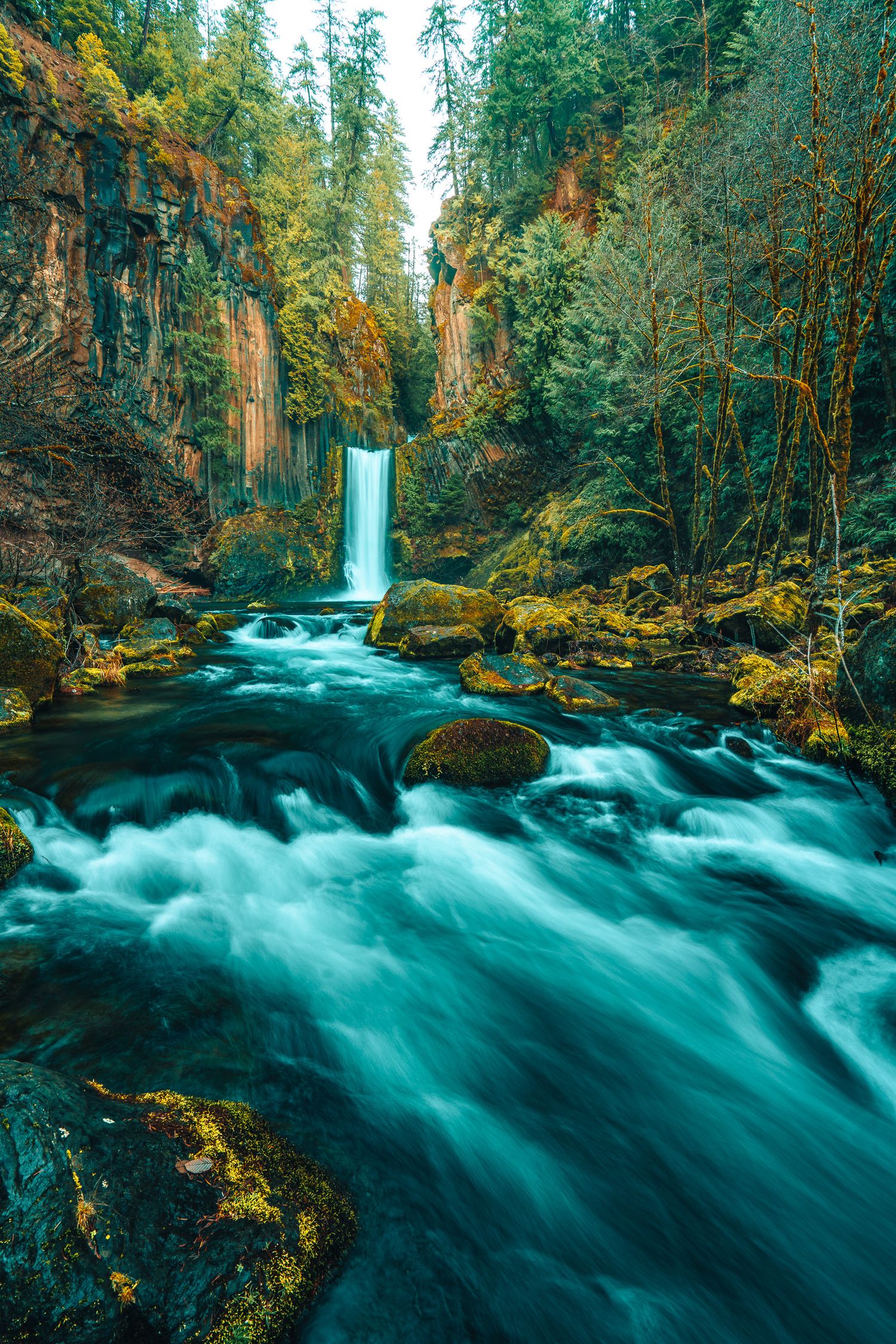 Powerful waterfall cascading into a rushing turquoise river surrounded by mossy rocks and dense forest.