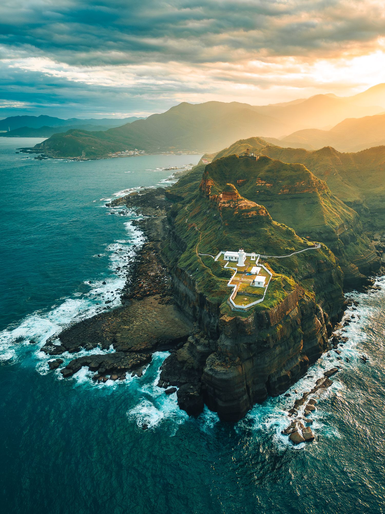 Aerial view of a dramatic green coastal cliff with white buildings overlooking crashing ocean waves at sunset.