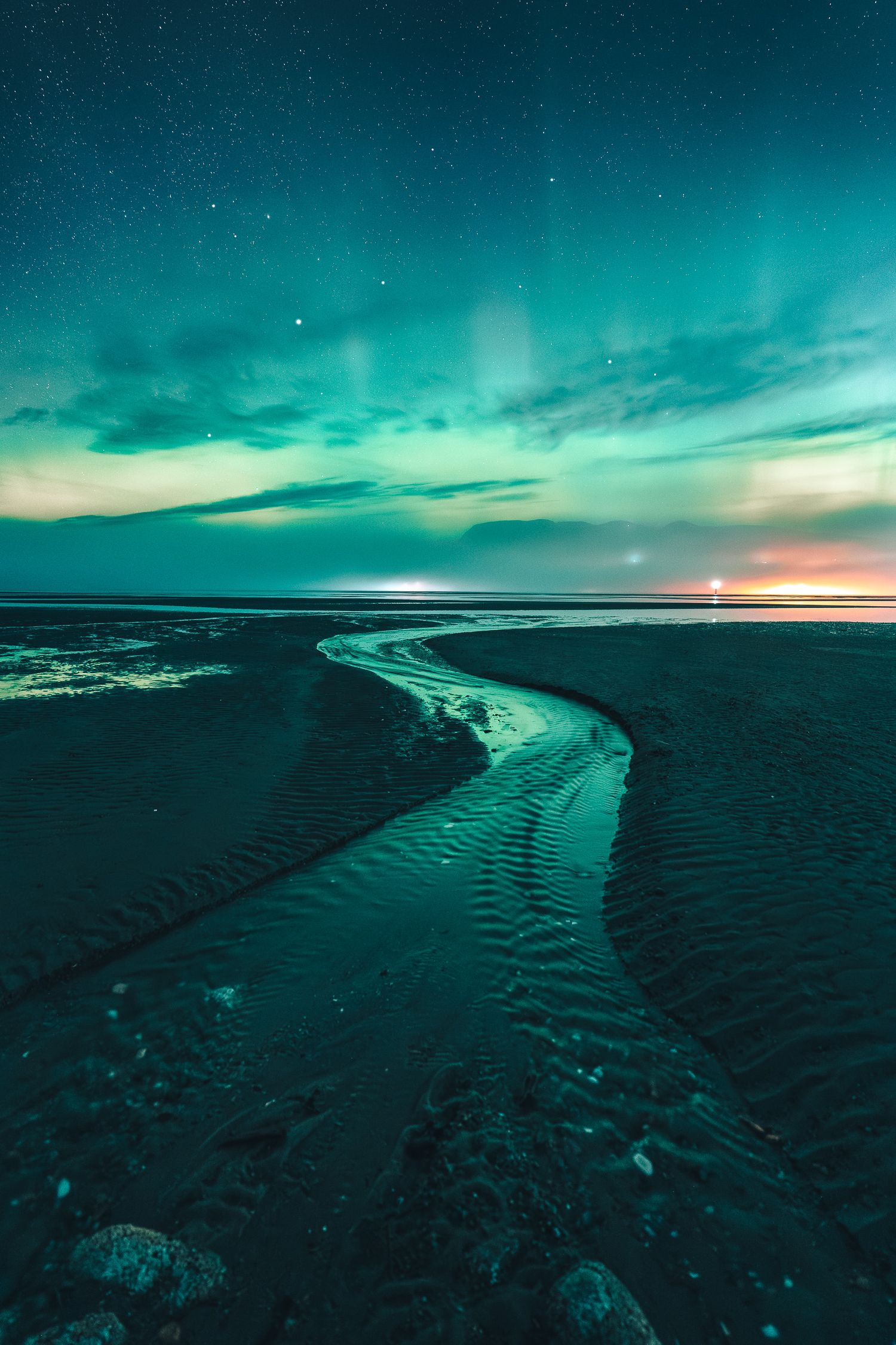 Aurora borealis glows in teal and green over a winding tidal stream on a dark sandy beach at night.