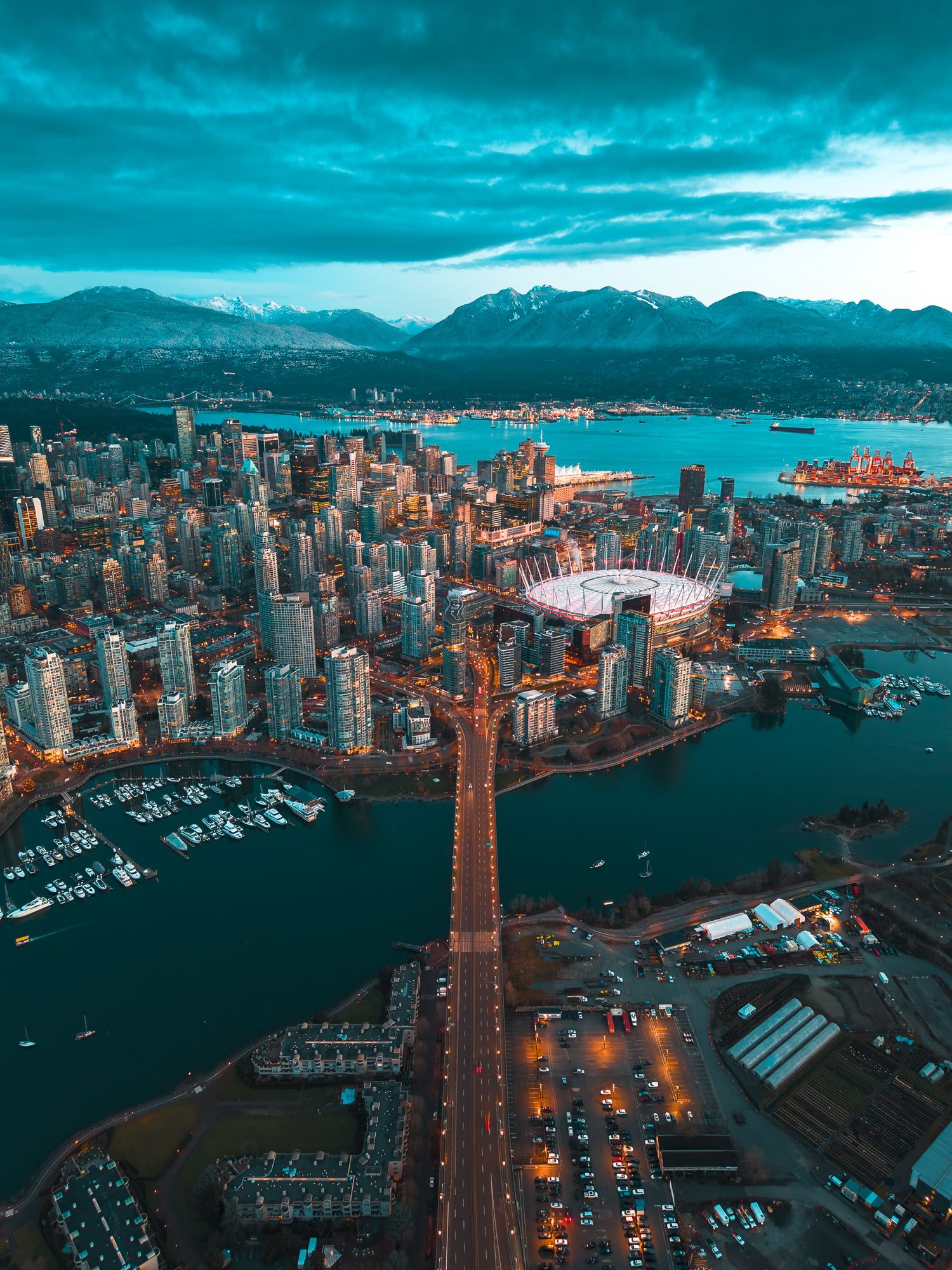 Aerial dusk view of Vancouver skyline with BC Place stadium, mountains, and illuminated bridges.