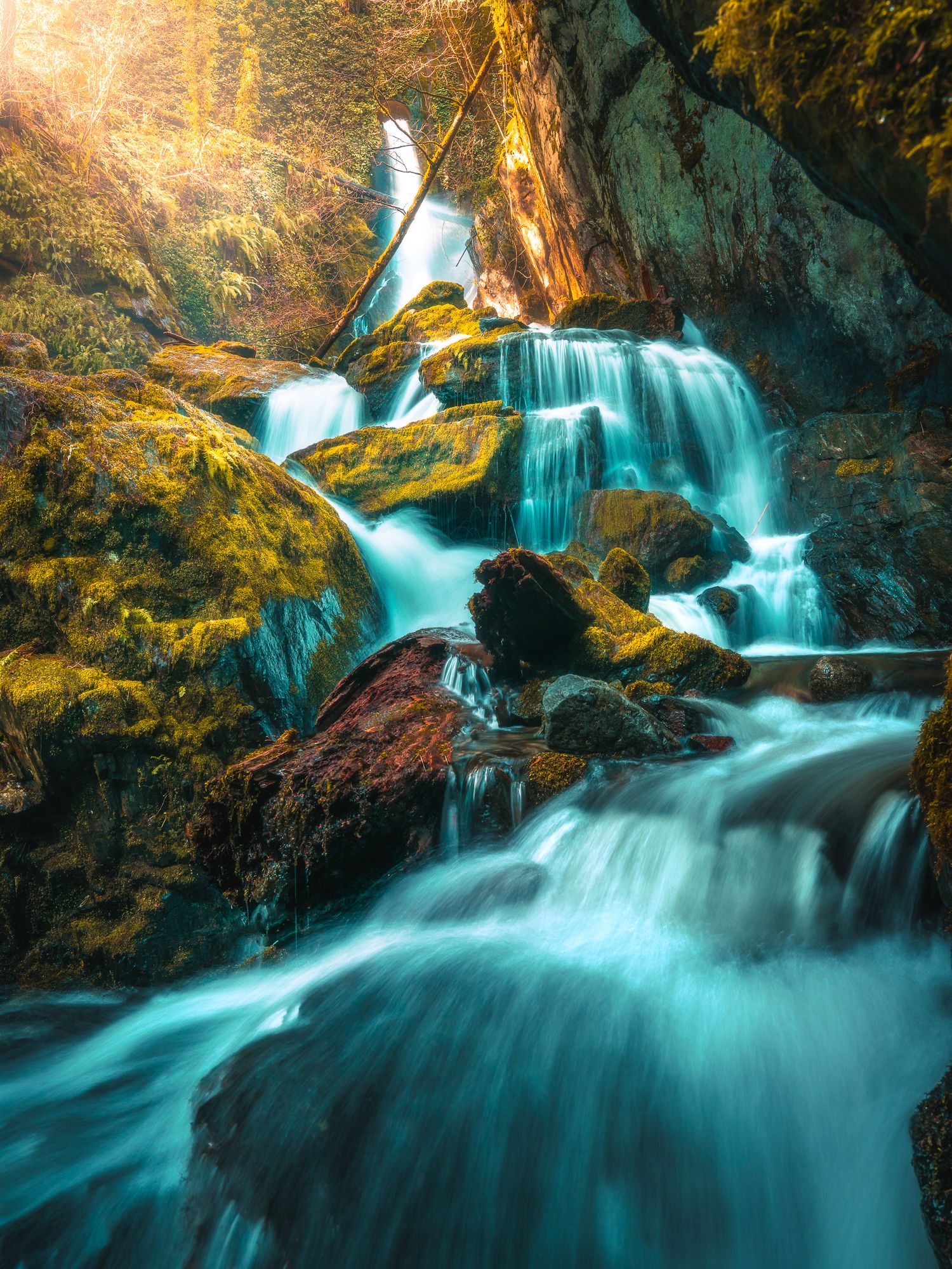 Turquoise waterfall cascading over moss-covered rocks in a sunlit forest grotto.