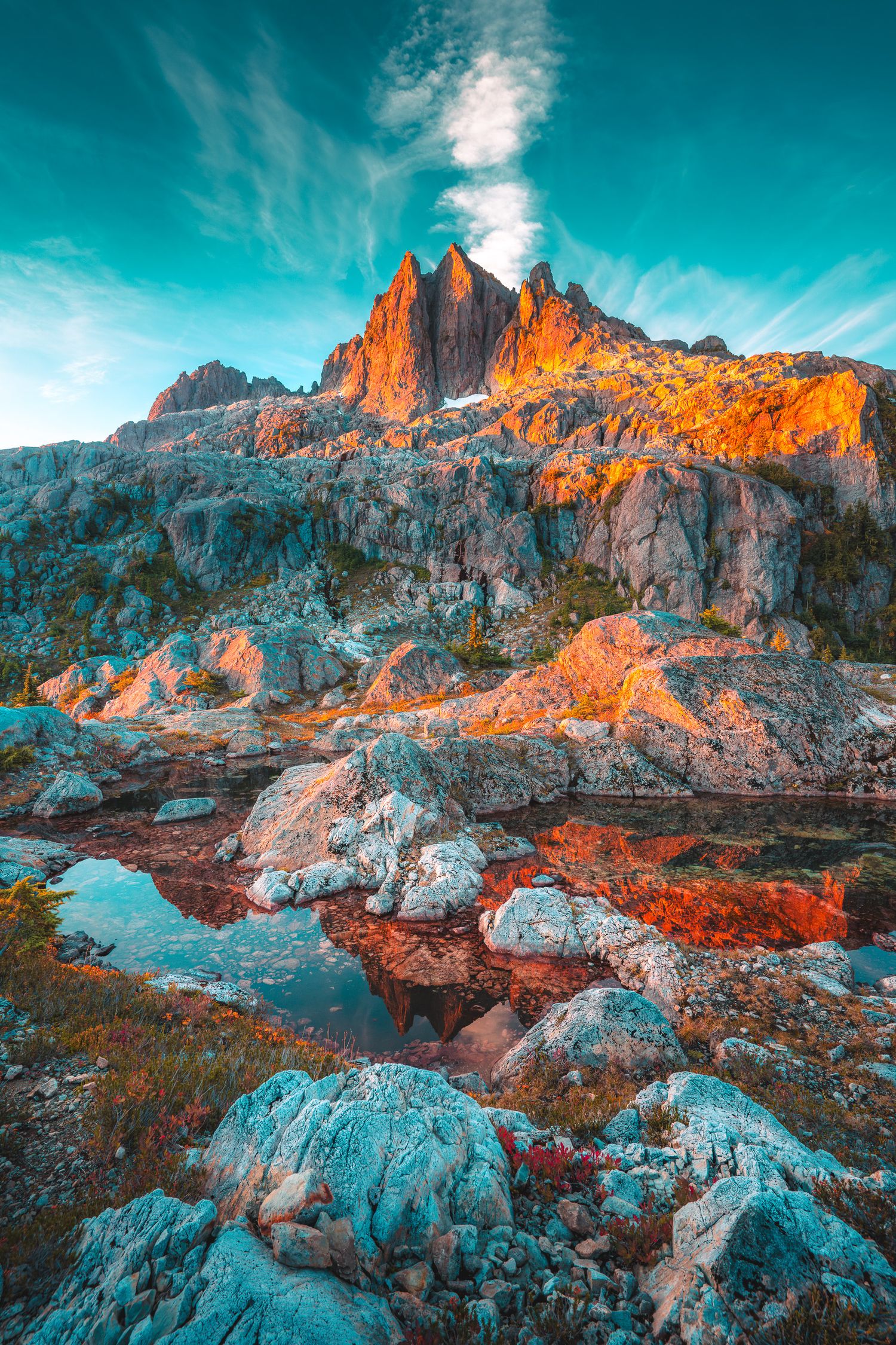 Dramatic orange-lit mountain peak reflected in a small alpine pool surrounded by rocky teal-toned terrain.