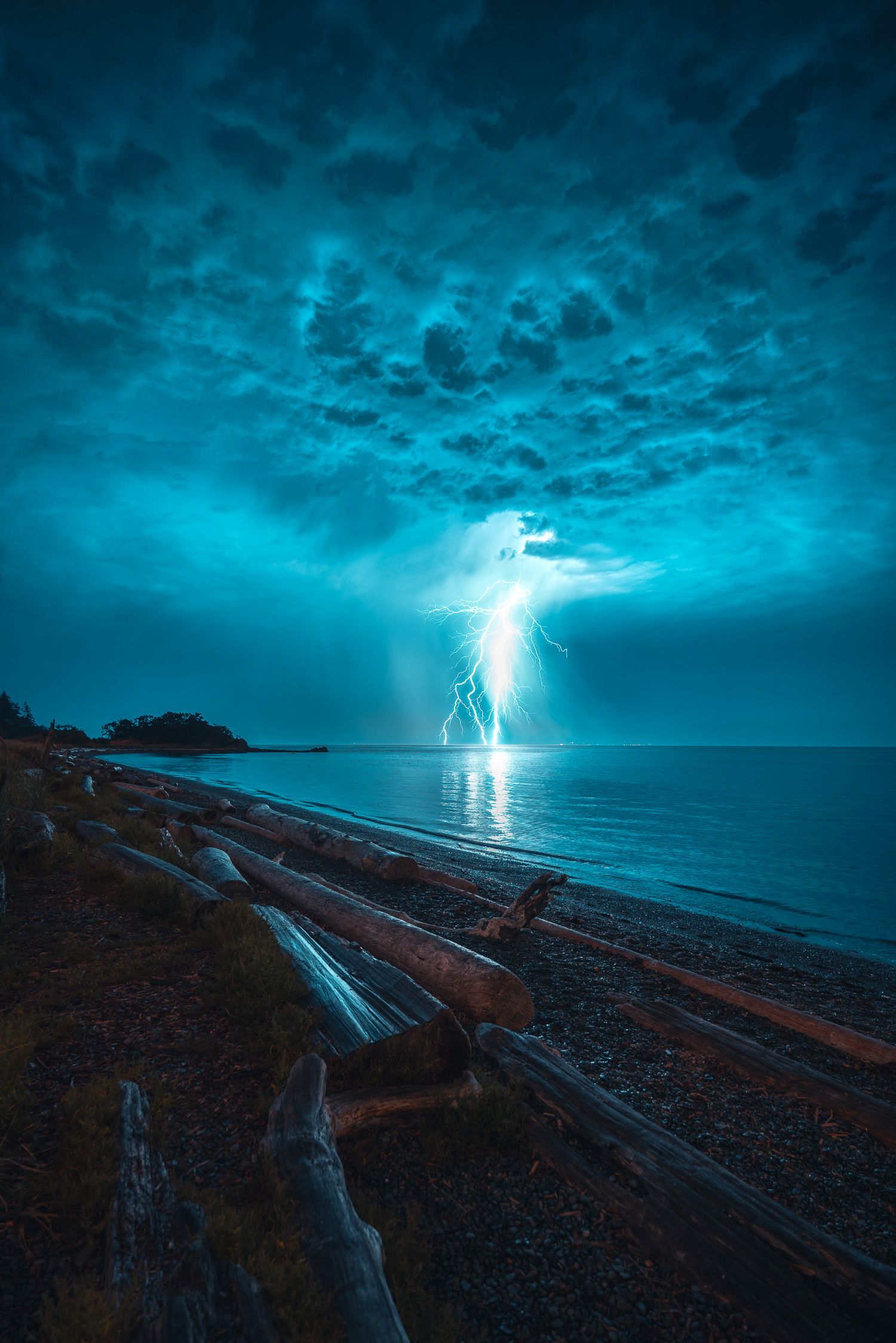Lightning strikes over a calm ocean during a dramatic storm, illuminating dark blue clouds above a driftwood-lined beach.