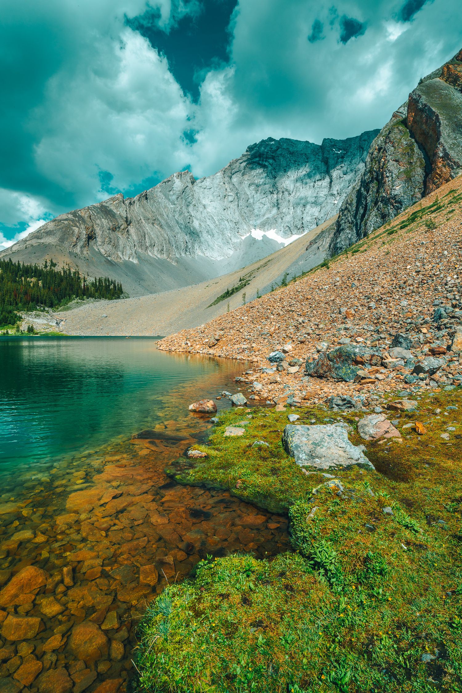 Crystal clear turquoise alpine lake with rocky shoreline and snow-capped mountain peaks under cloudy skies.