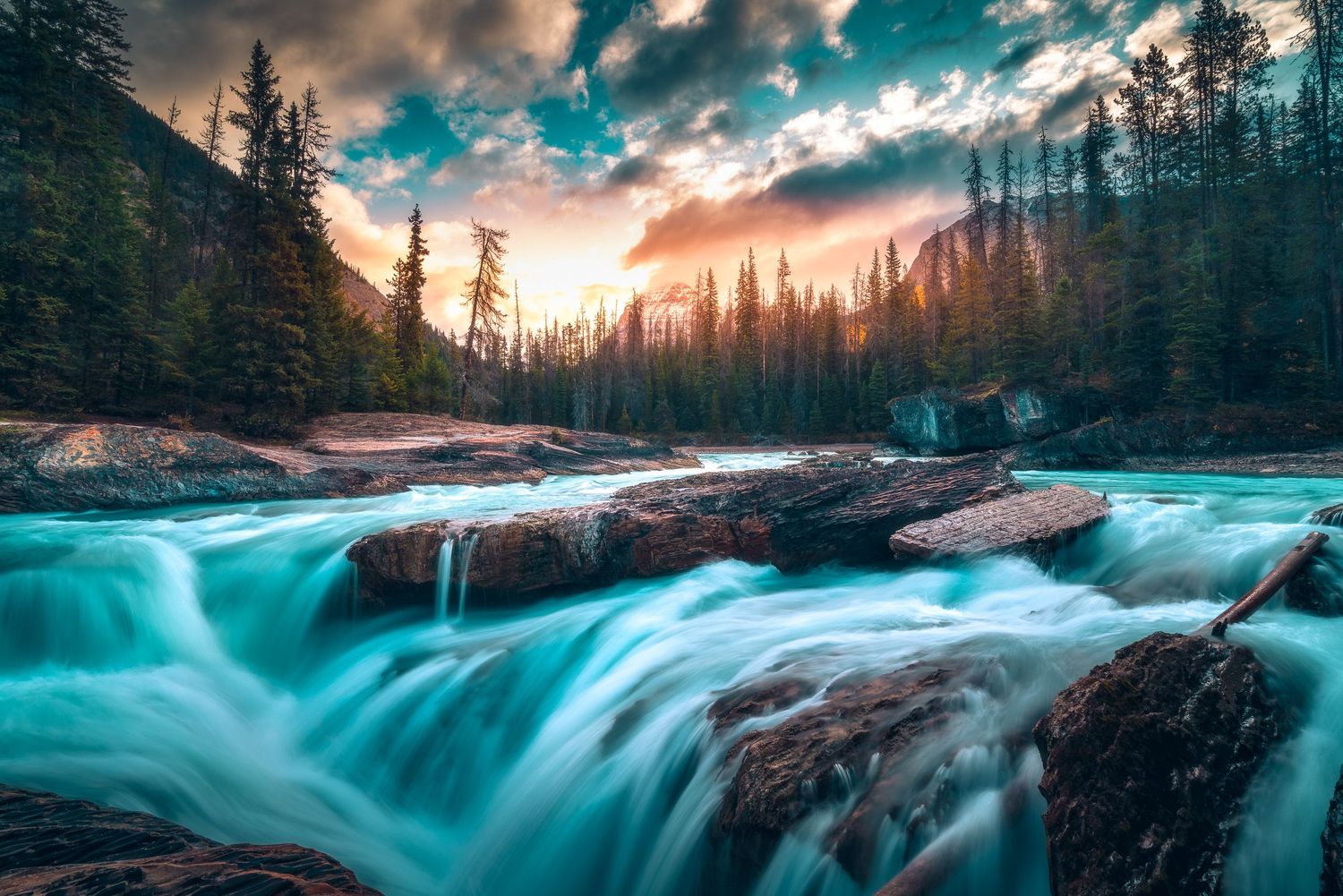 Turquoise waterfall cascading over rocks in a forest at dramatic sunset with glowing clouds.