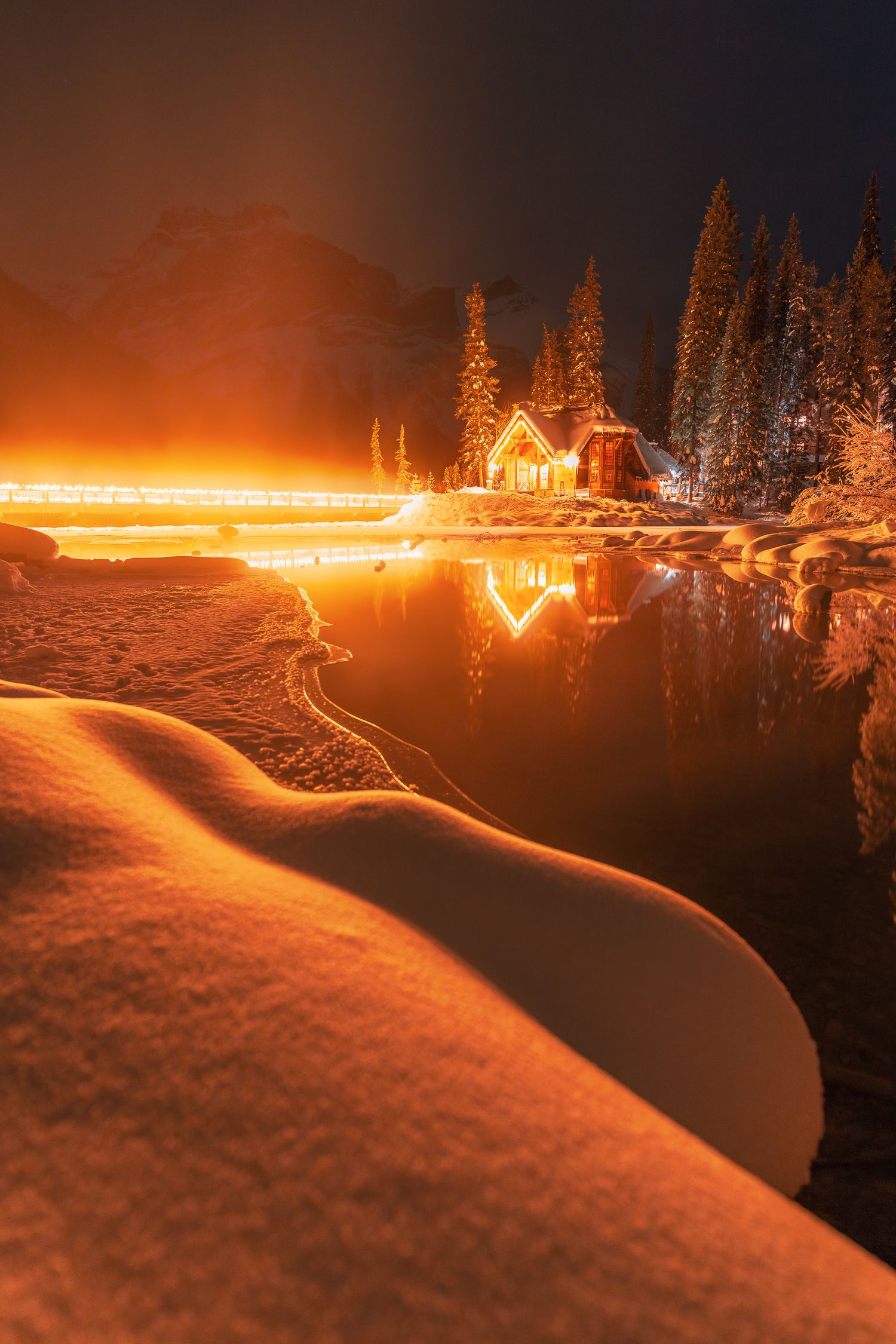 Cozy illuminated cabin reflects warmly on a snowy winter lake at night in the mountains.