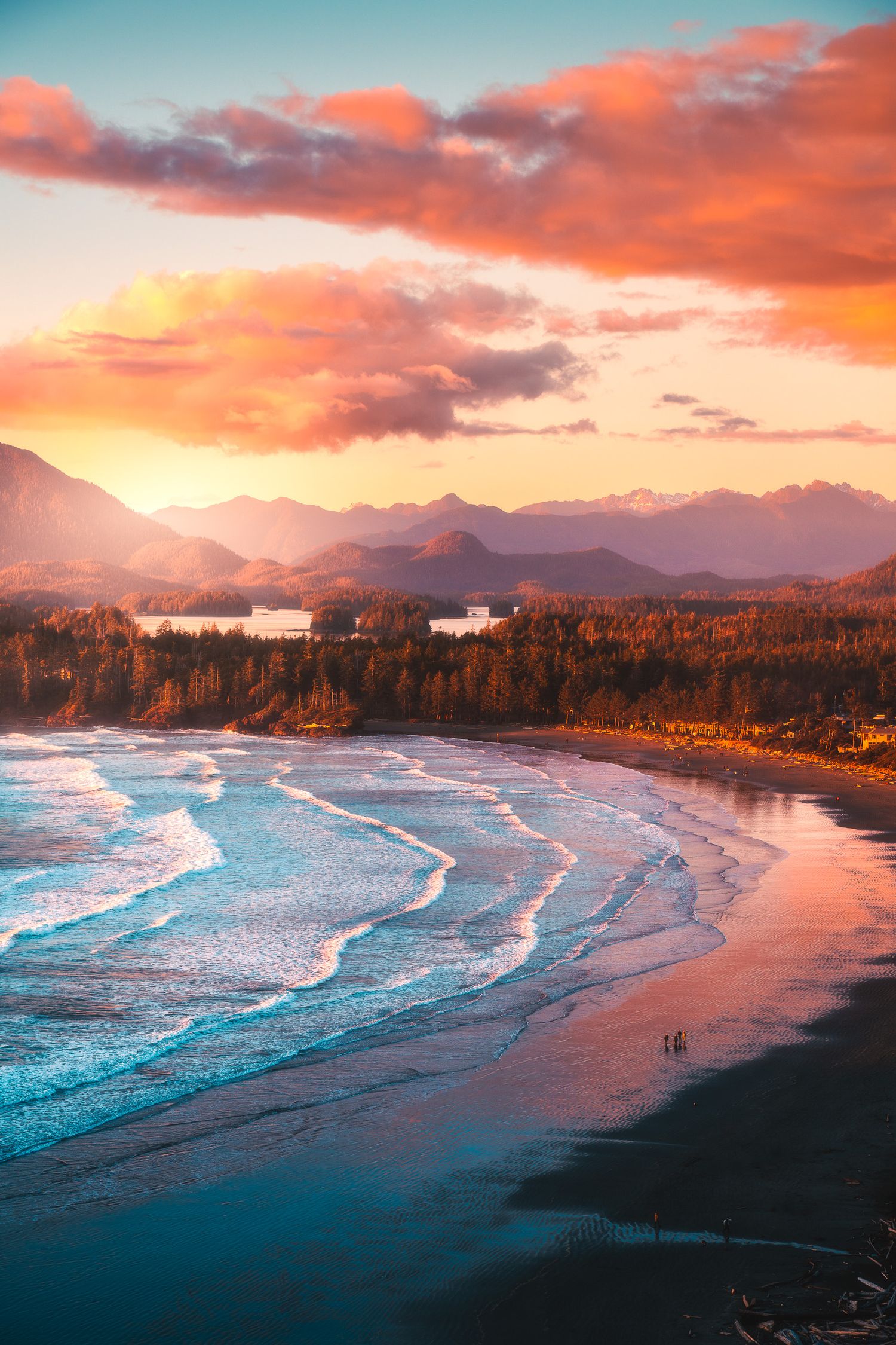 Stunning sunset over coastal beach with golden waves, forested shore, and mountain backdrop.