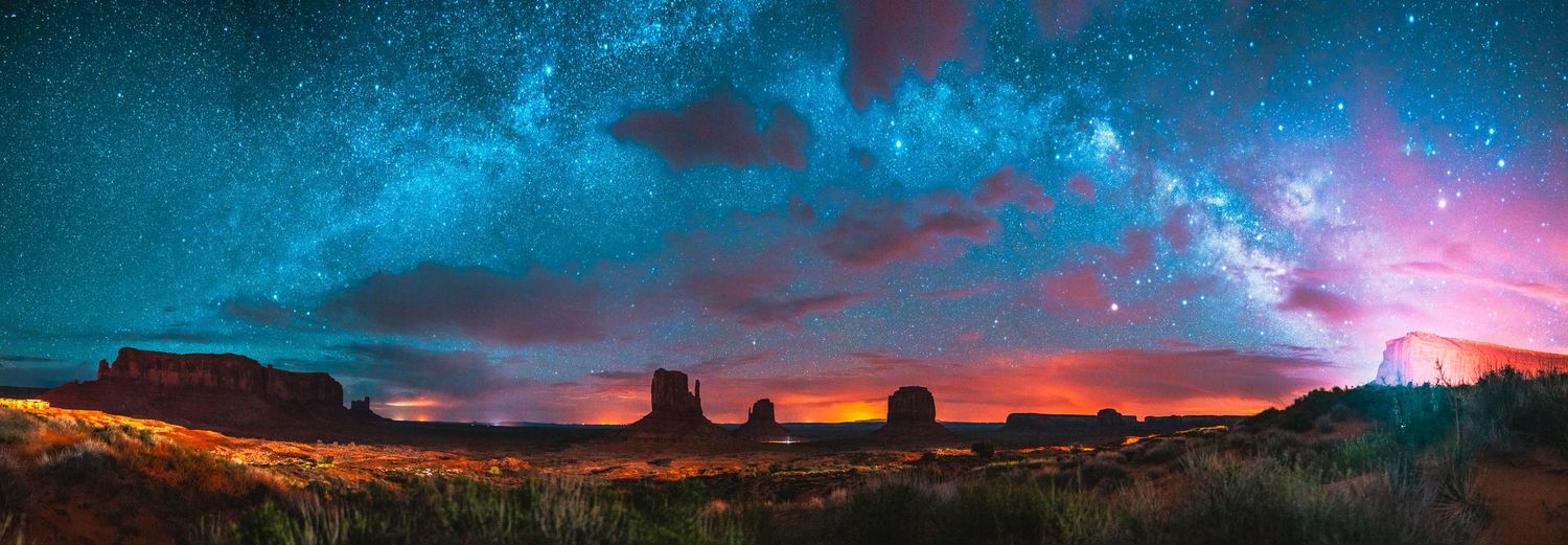 Panoramic night sky with Milky Way over Monument Valley's iconic sandstone buttes glowing under a starlit desert horizon.