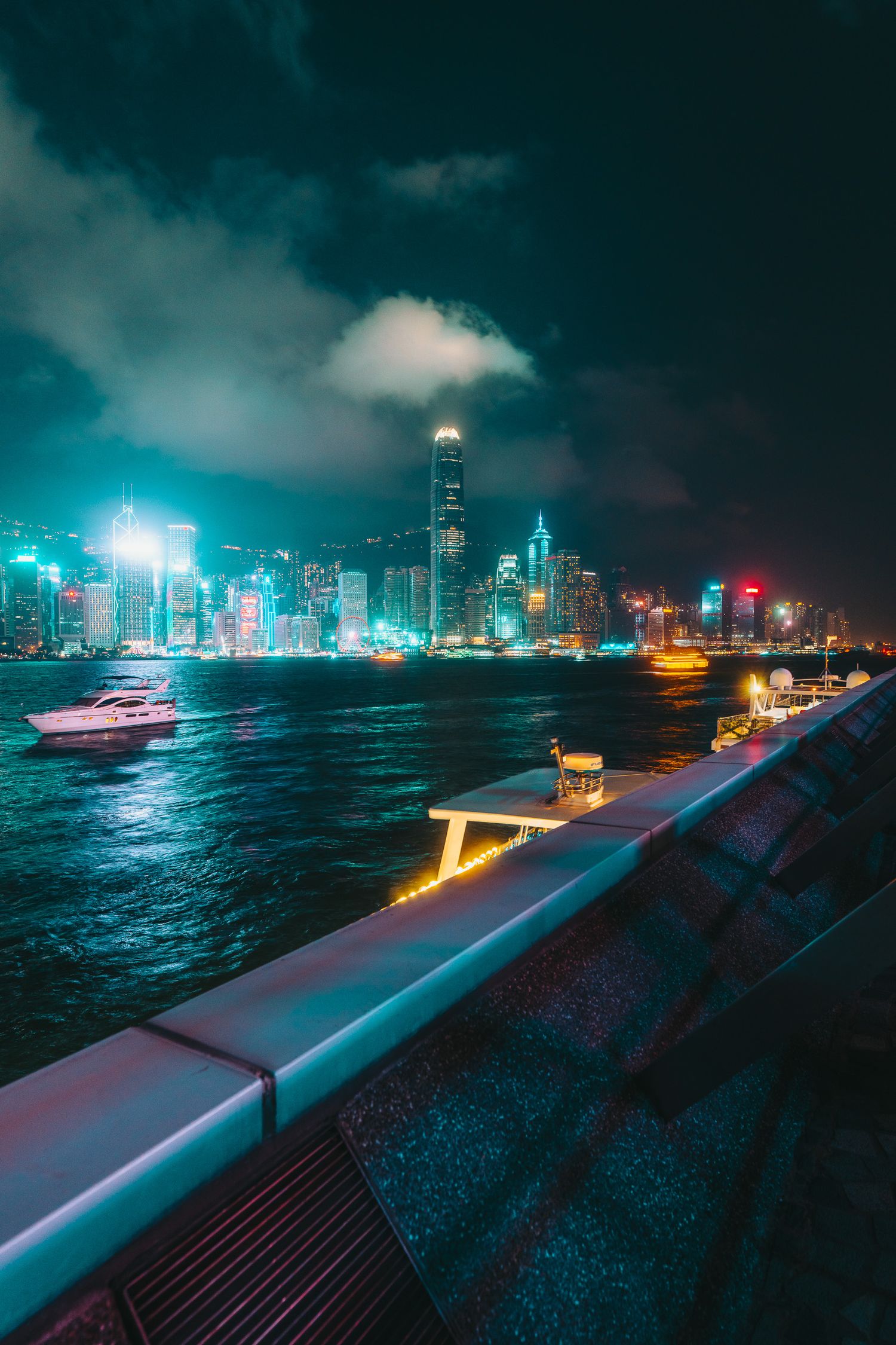 Hong Kong's iconic illuminated skyline reflects on Victoria Harbour waters viewed from a waterfront promenade at night.