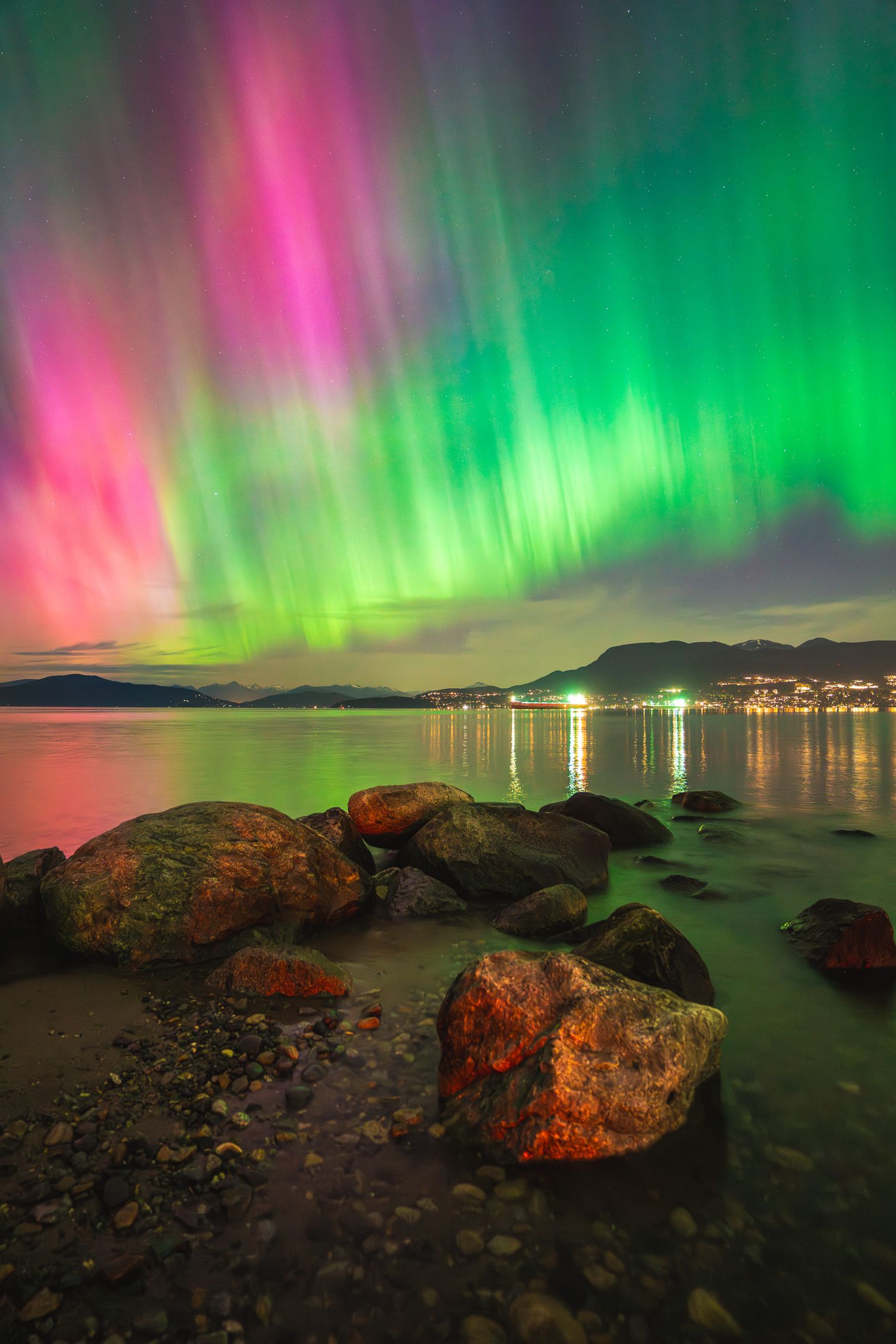 Dramatic pink and green northern lights aurora borealis illuminate a rocky shoreline at night.