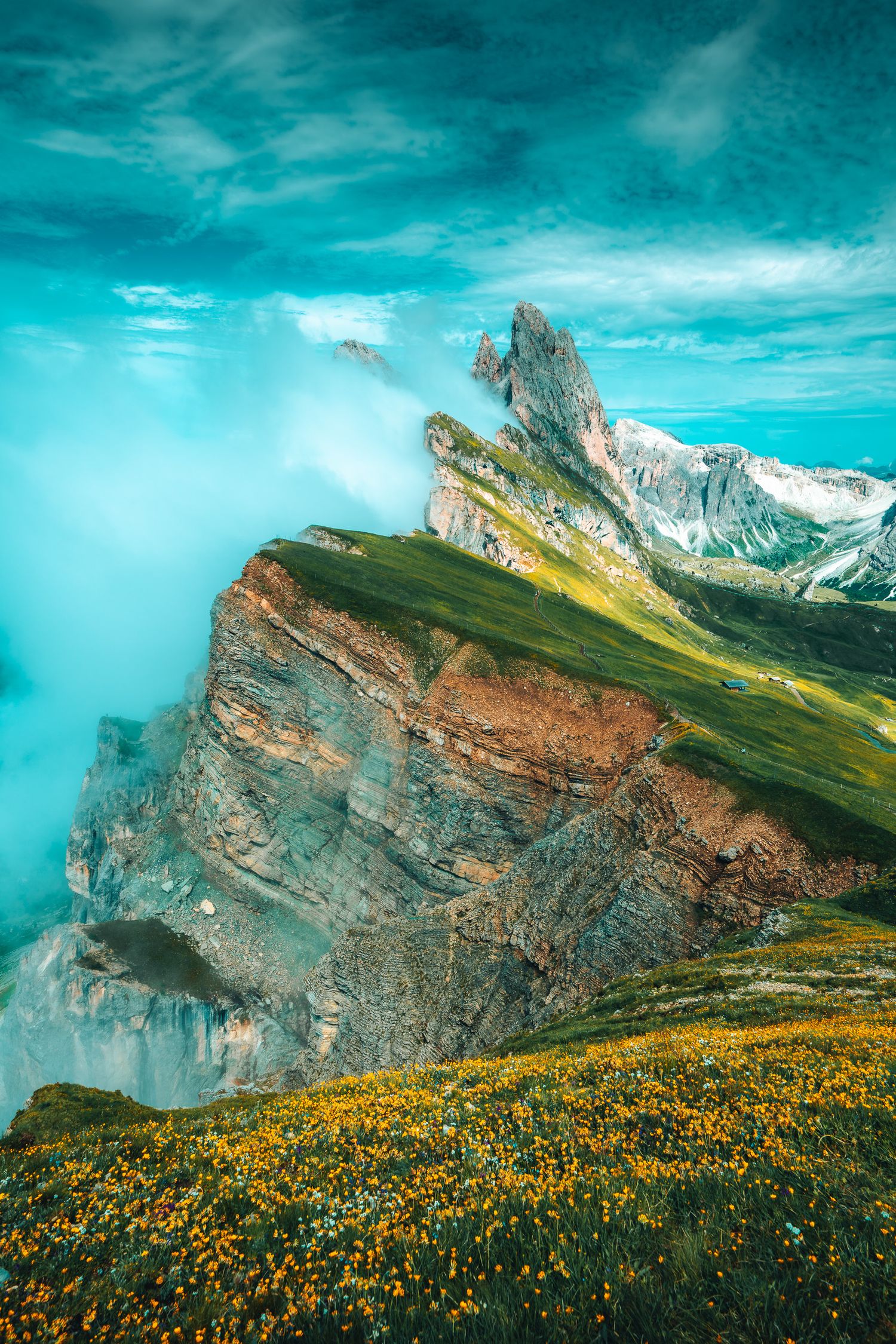 Dramatic Dolomite mountain cliffs with yellow wildflowers, green slopes, and misty turquoise sky in the Italian Alps.