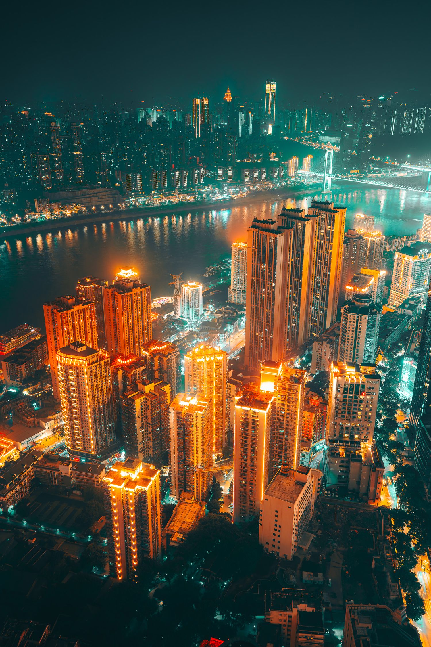 Aerial night view of Chongqing's golden-lit skyscrapers along a winding river with glowing city lights below.